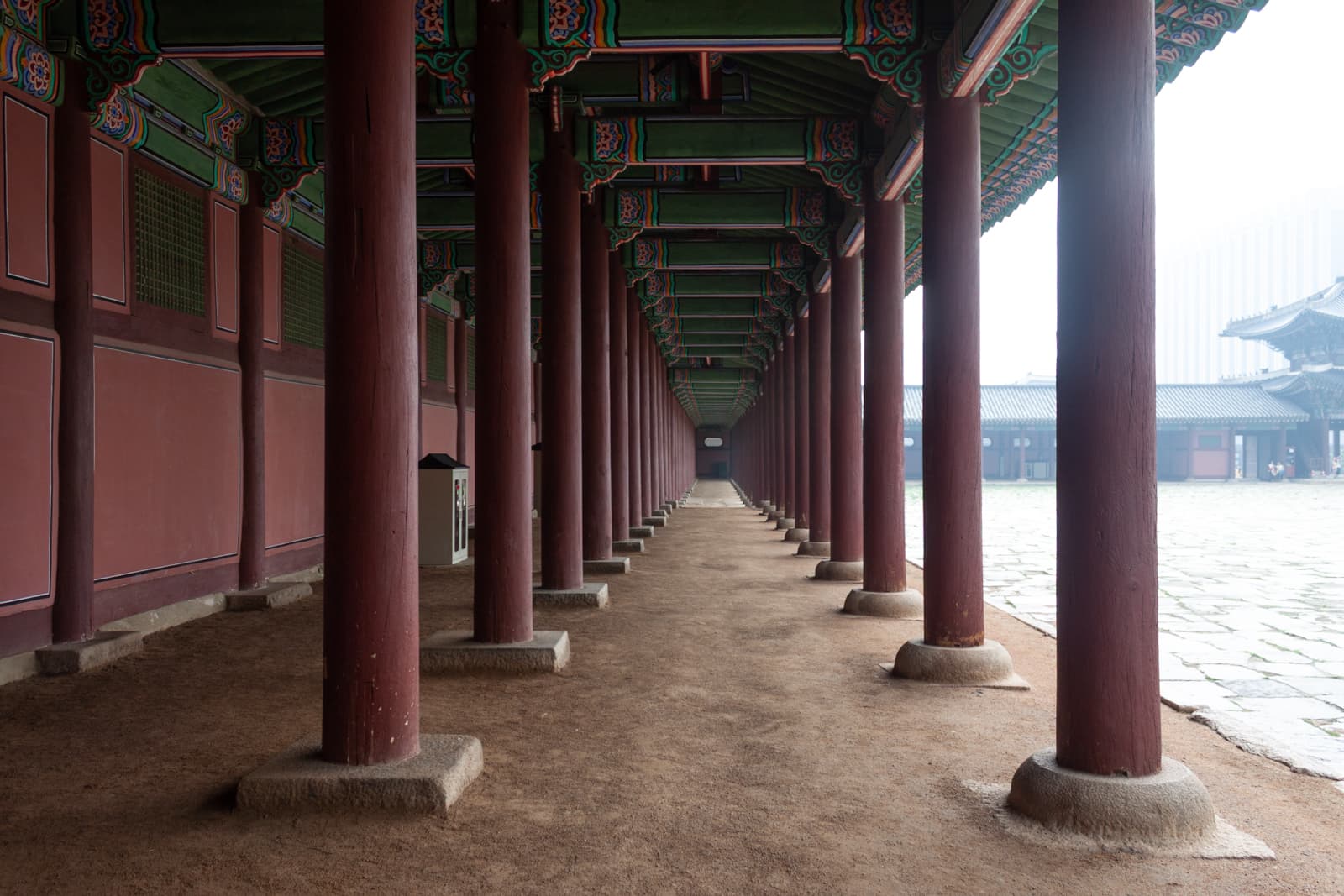 A long corridor of a traditional Korean palace with red columns supporting a beautifully decorated ceiling. The path is made of dirt, extending into the distance, creating a sense of symmetry and depth.