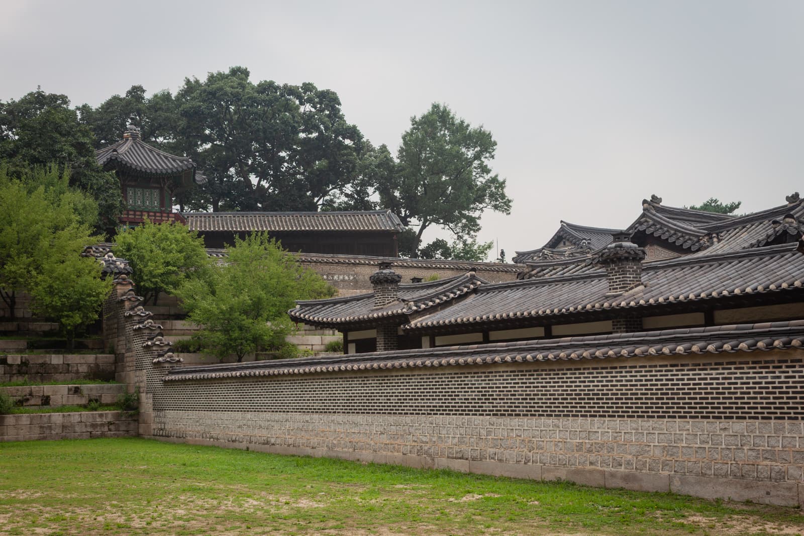 A traditional Korean palace courtyard, featuring a series of tiled roofs and brick walls. Trees and stone steps line the background, blending nature with the historical architecture.