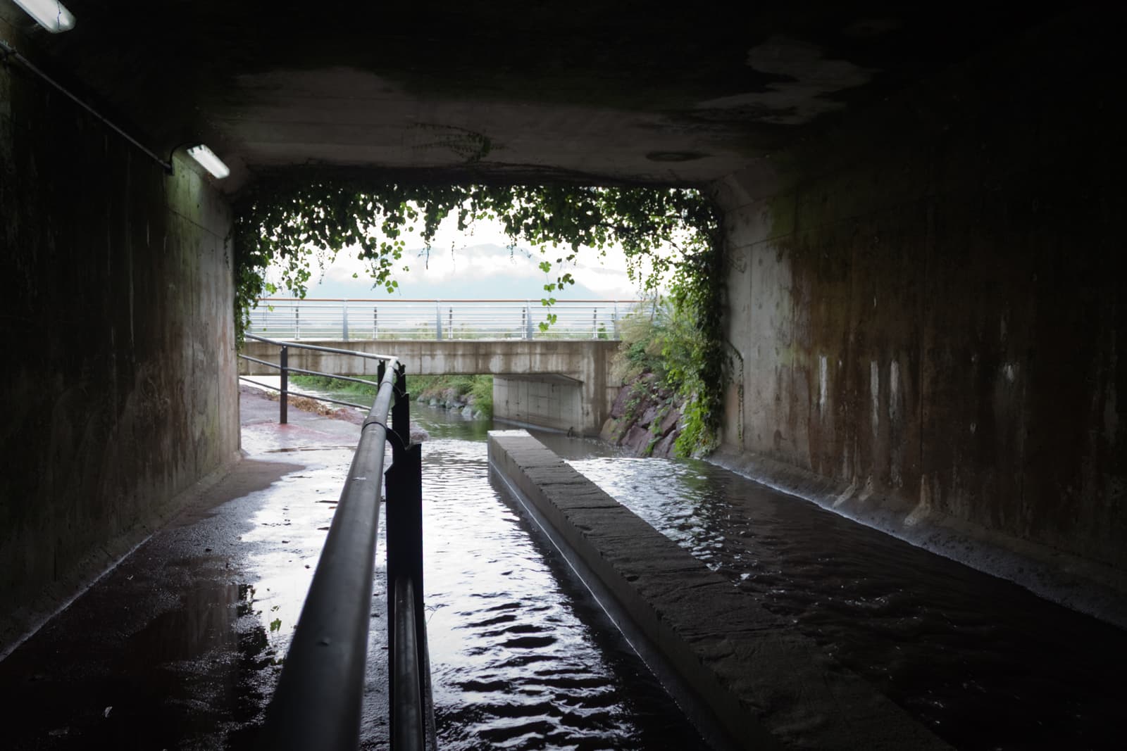 A view from a tunnel under a bridge. Sunlight illuminates the path, with water flowing through the channel. Vines hang from the top of the tunnel, framing the view of the bridge and the mountains beyond.