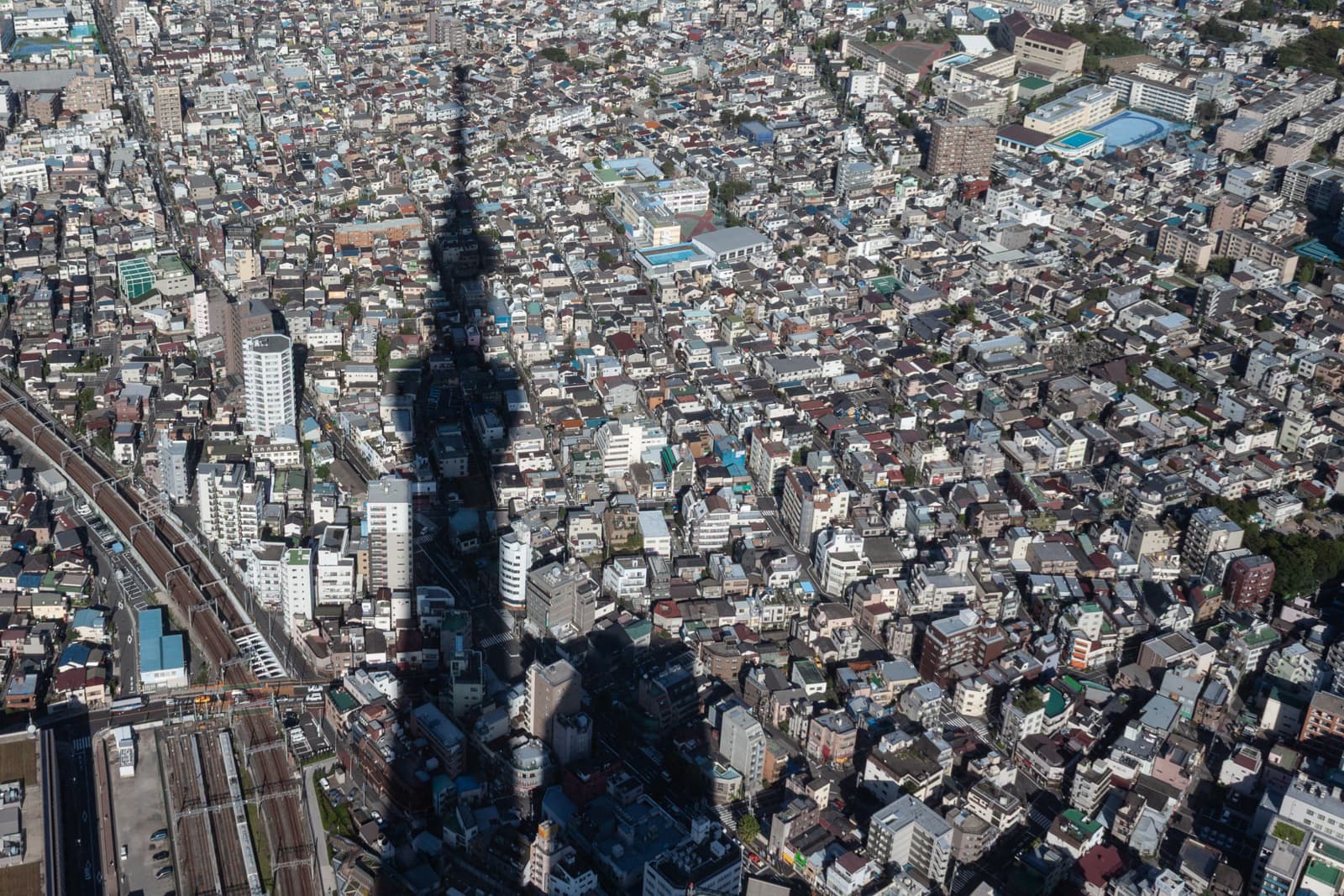 Aerial view of a densely packed urban city with buildings of various sizes. The shadow of a tall structure, possibly a tower, stretches across the cityscape, creating a dramatic contrast against the clustered buildings below.