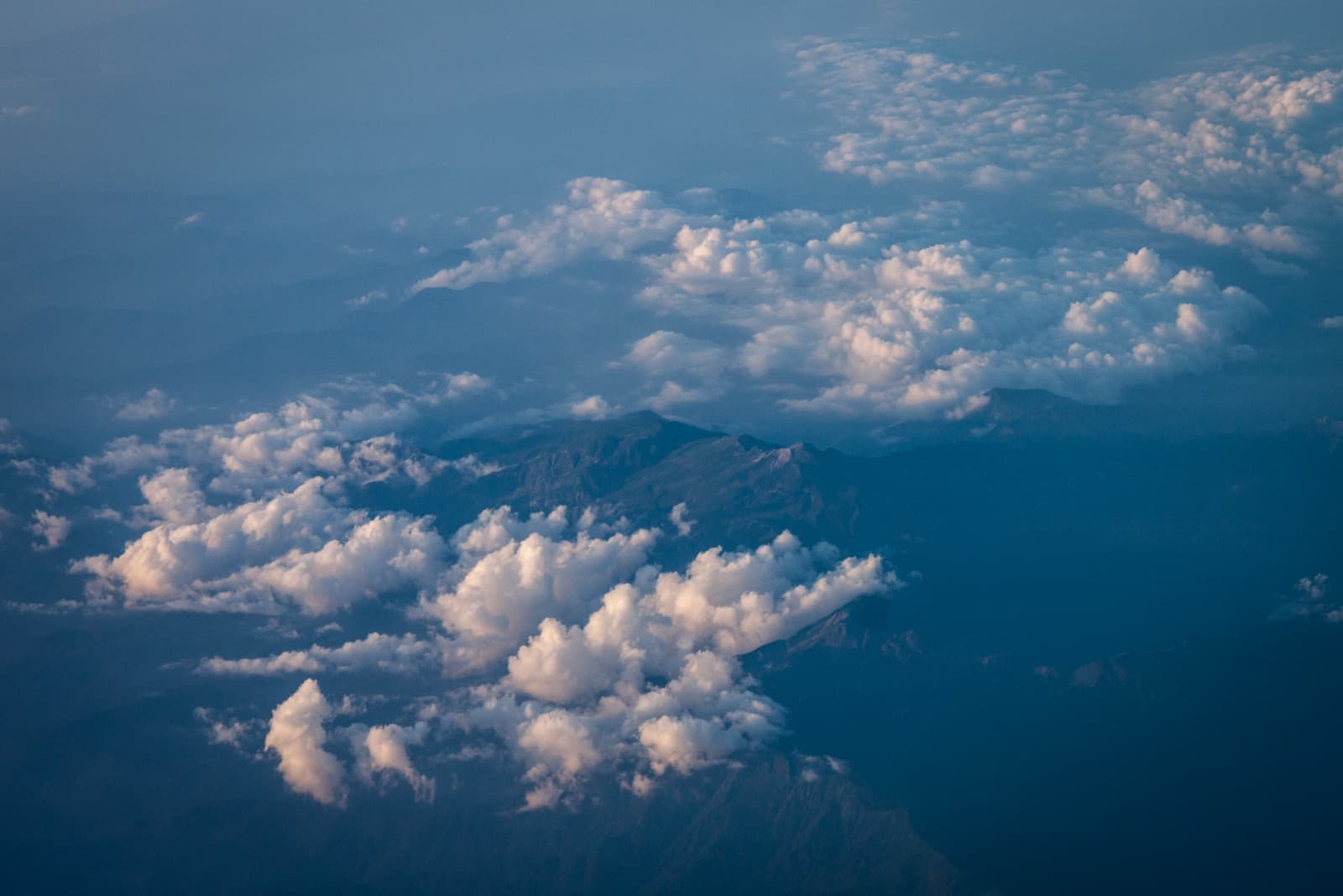 A high-altitude view of a mountainous landscape partially obscured by clouds. Puffy white clouds float above the rugged terrain, with mountain peaks visible through breaks in the cloud cover, creating a serene and expansive atmosphere.