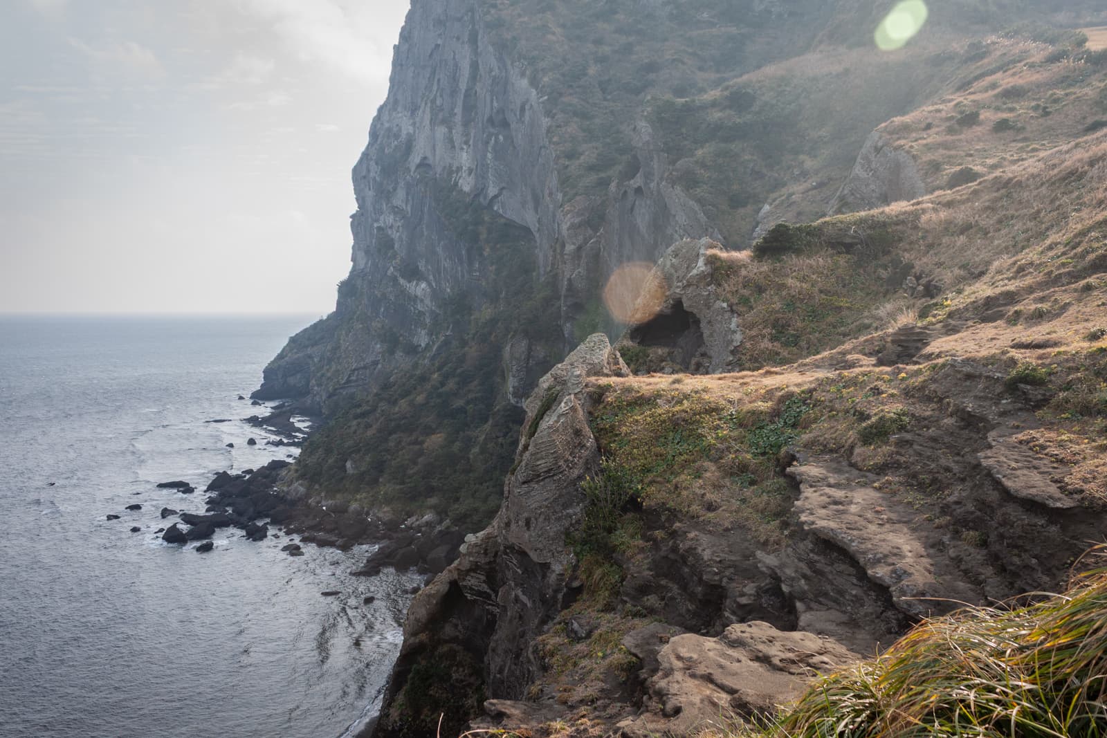 A dramatic coastal cliffside with a rugged path leading up the steep incline. The cliff drops off into the ocean below, where waves crash against the rocks, while patches of sunlight filter through the cloudy sky, casting a soft glow on the landscape.