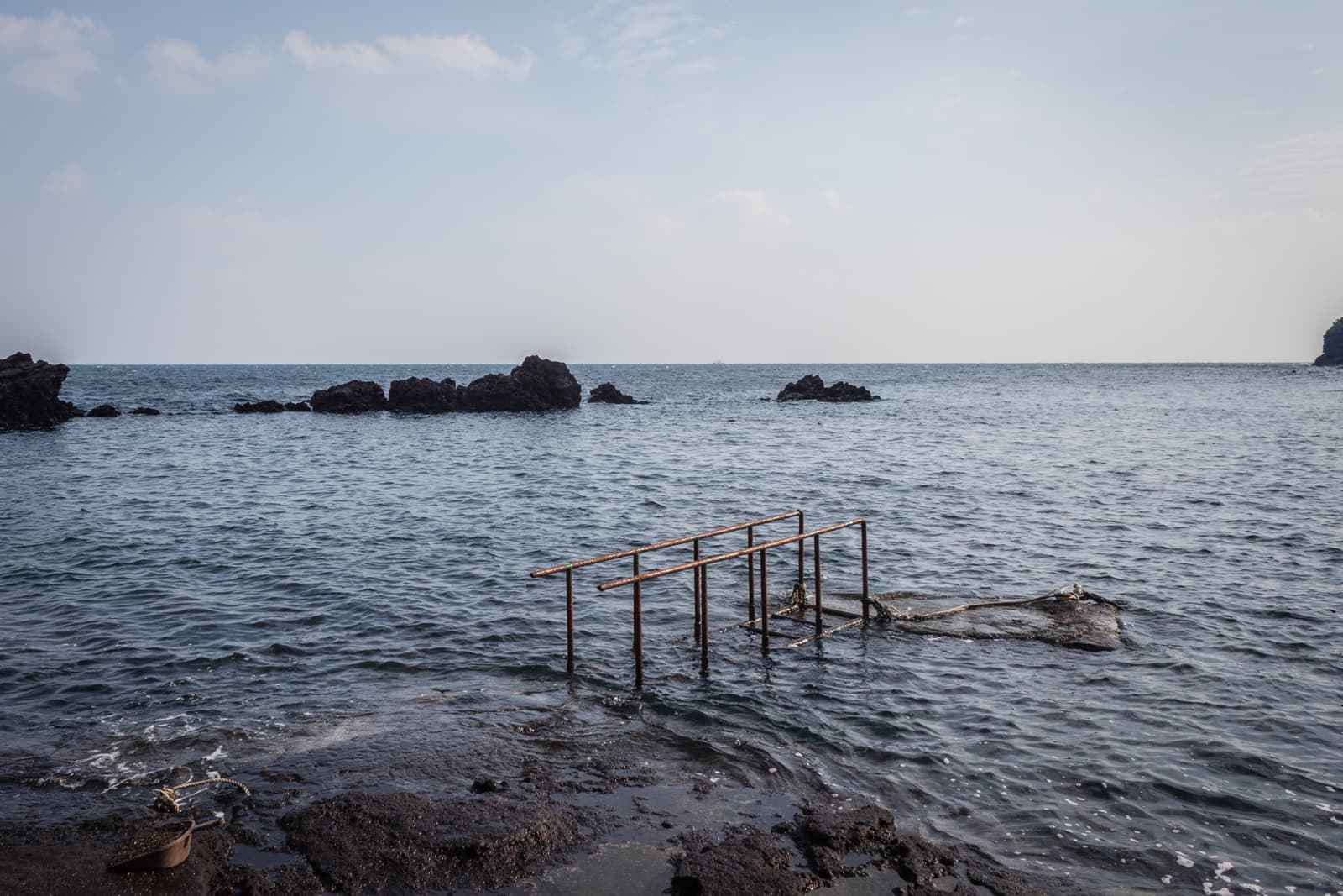 A rocky seashore with an old, rusted handrail extending into the shallow water. The calm sea stretches out toward distant rocks, while the overcast sky creates a serene, contemplative atmosphere.