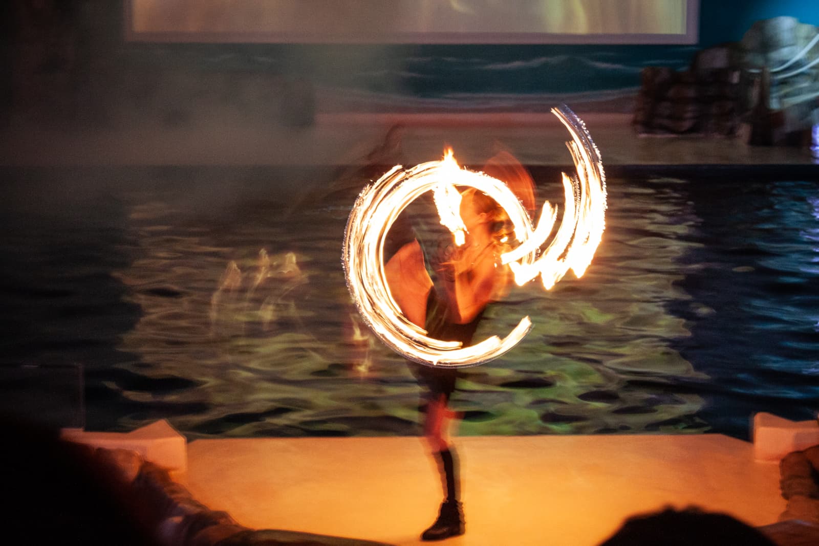 A performer spins flaming torches, creating swirling patterns of fire in a darkened setting. The movement and light blur the figure, capturing the dynamic energy of the fire dance.