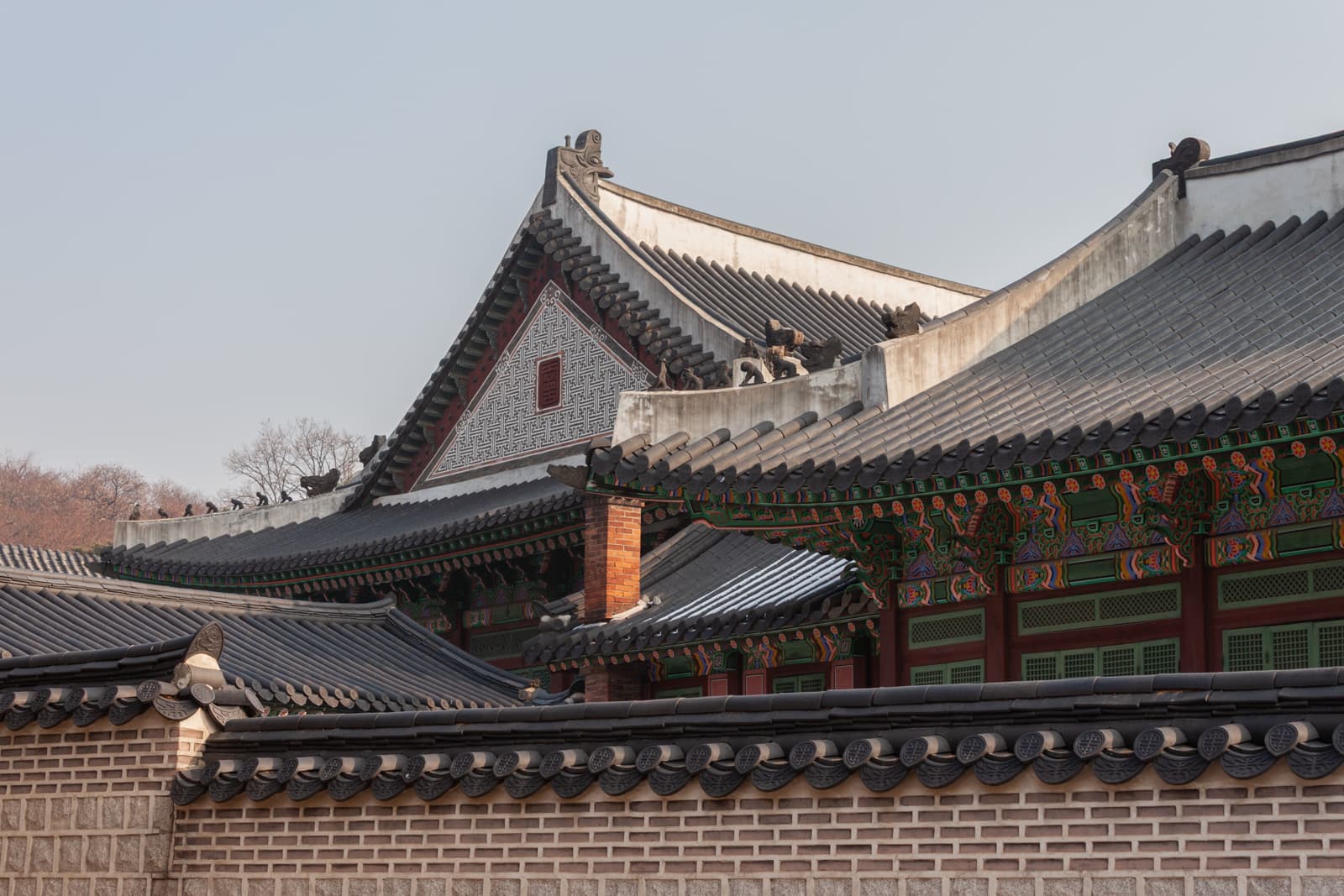 A complex of traditional temple rooftops with intricate eaves, painted with colorful patterns, is visible against a pale sky. The walls are tiled, and the architecture showcases historical design elements.
