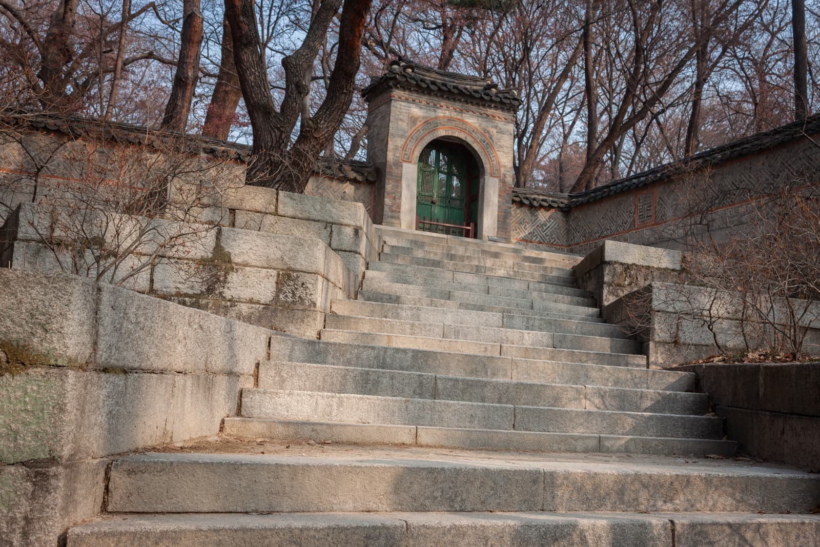 A steep stone stairway leads up to a traditional arched gate set within an ancient stone wall. Trees with bare branches frame the steps on either side.