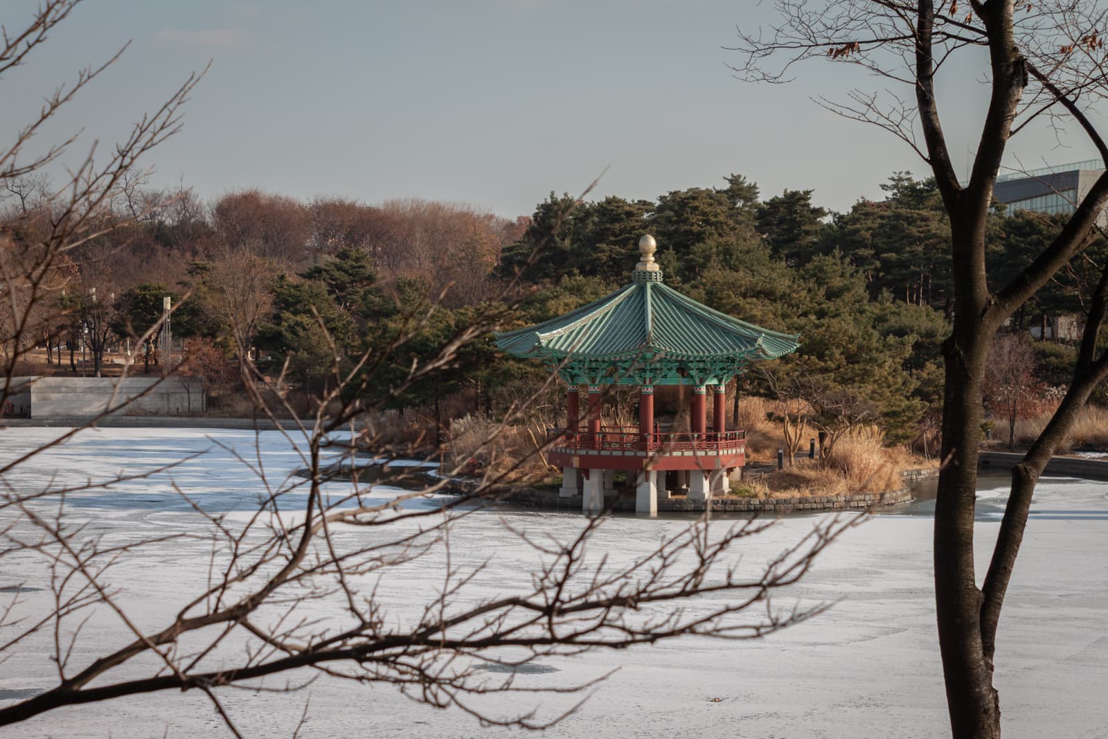 A small, ornate gazebo with a green, pointed roof stands beside a frozen pond. Trees with bare branches frame the scene, while the gazebo overlooks a serene winter landscape.