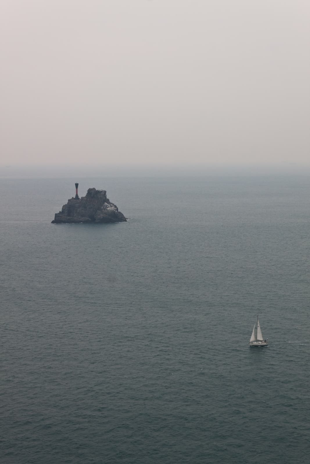 A lone sailboat navigating across a calm, expansive ocean, with a small rocky island featuring a prominent beacon in the distance. The sky appears overcast, casting a muted tone over the scene.