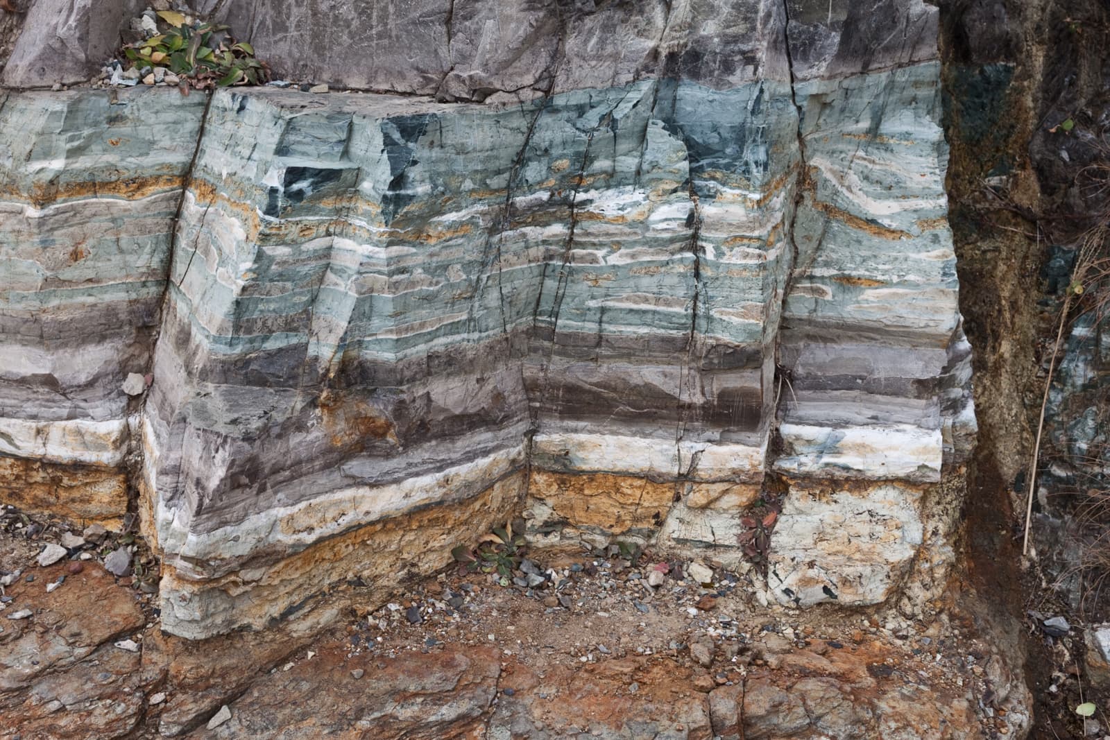 Close-up view of stratified rock layers in varying shades of green, brown, and white, showing the geological complexity of the cliffside. The layers appear weathered and cracked in places.