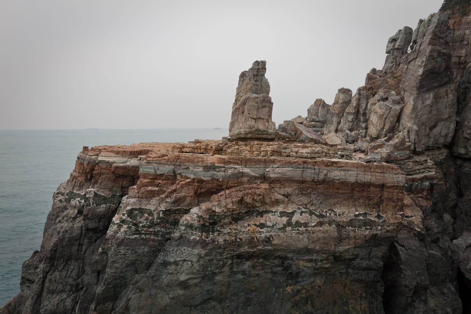 A rugged, towering rock formation standing along the coastline, with dramatic layered cliffs stretching toward the sea. The sky is overcast, and the rough textures of the rocks contrast with the smooth water below.