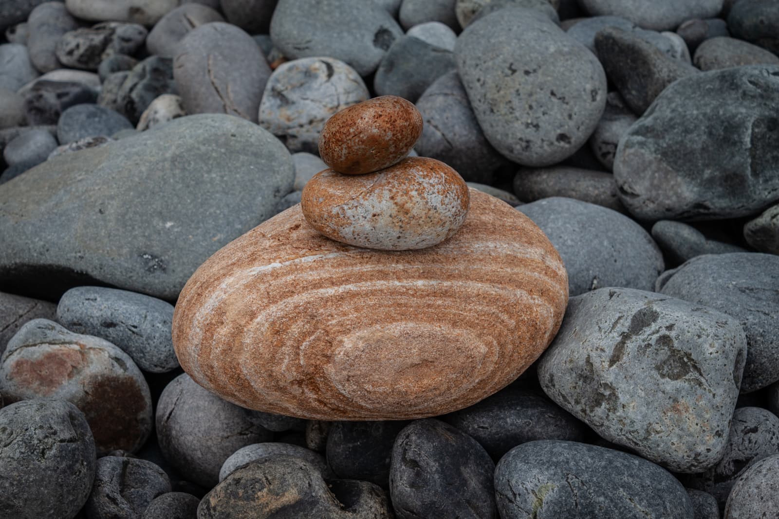 A balanced stack of smooth, rounded stones placed on a bed of gray pebbles. The central stone has concentric patterns, while smaller stones rest atop it, creating a simple yet natural sculpture.