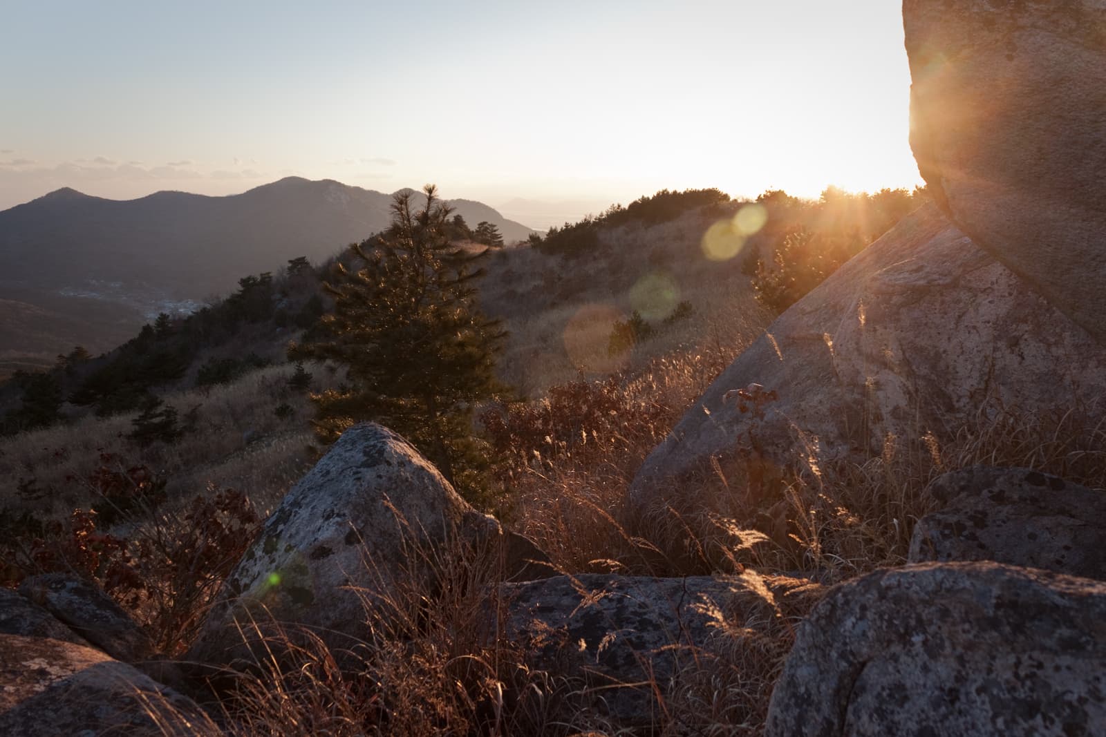 Sunlight filtering through the mountains, casting a warm glow on rocks and trees, creating a serene landscape at sunset.