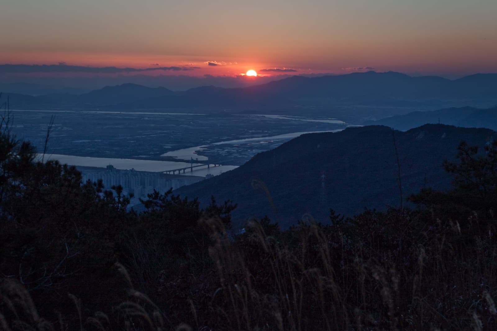 A stunning view of the sun setting behind mountains and a river winding through the valley, casting warm colors over the landscape.