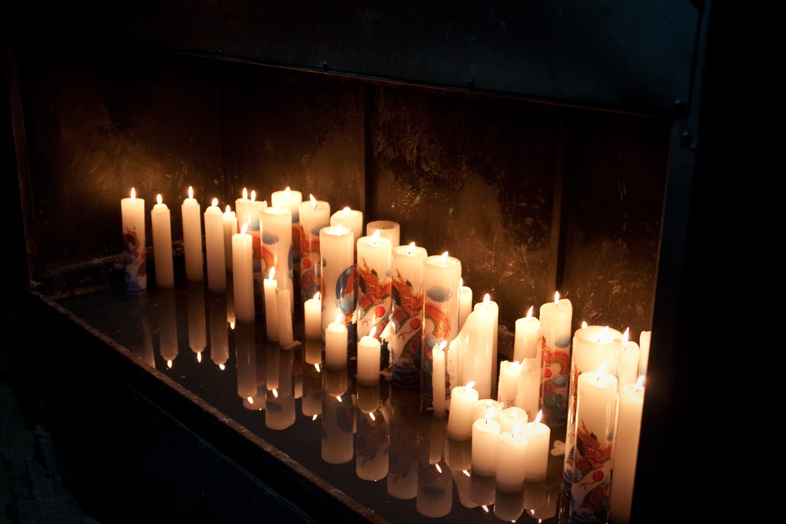 A group of candles lit in prayer, their flames reflected on a glossy surface, casting a warm and serene glow in the dark surroundings.