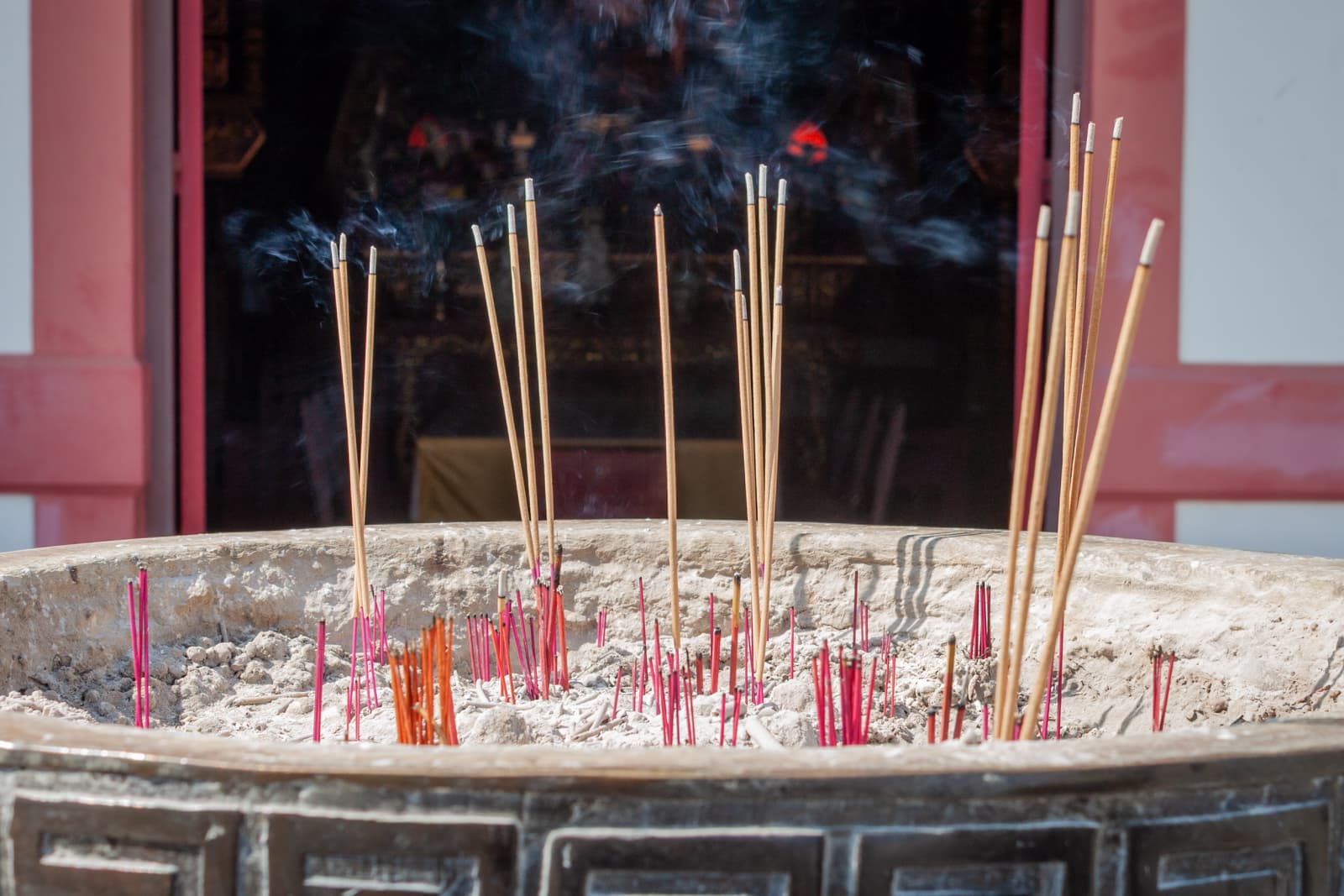 A large outdoor incense burner filled with thin, colorful incense sticks burning and releasing wisps of smoke. The scene is set in front of a temple, with light streaming in from one side.