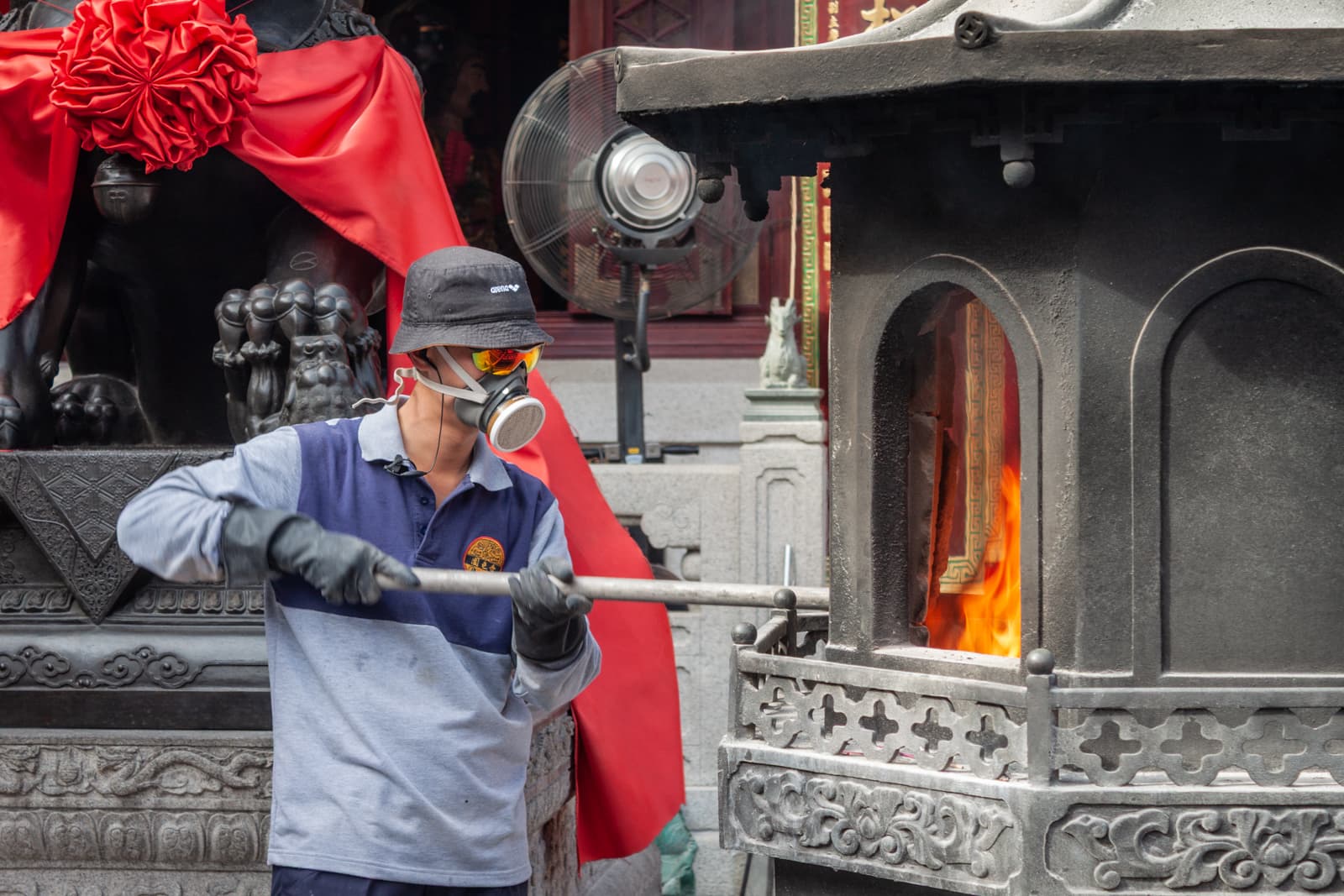 A man wearing protective gear, including a gas mask and gloves, tending to a fire inside a large incense burner at a temple. The surrounding area includes decorative statues and a bright red cloth draped around the entrance.