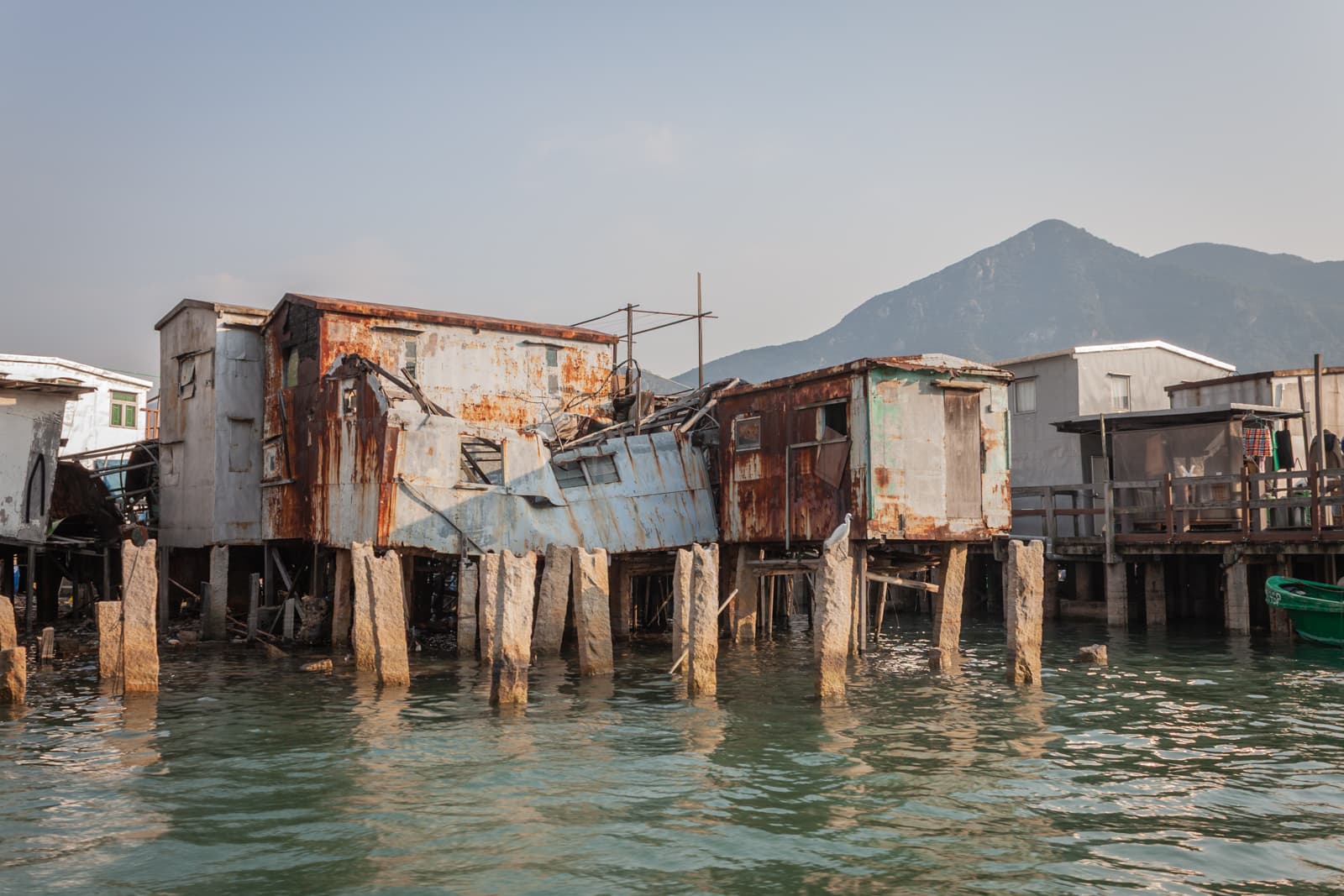 A pair of dilapidated wooden stilt houses, rusted and worn by time, standing above greenish water. The rugged mountains in the distance contrast with the aged structures.