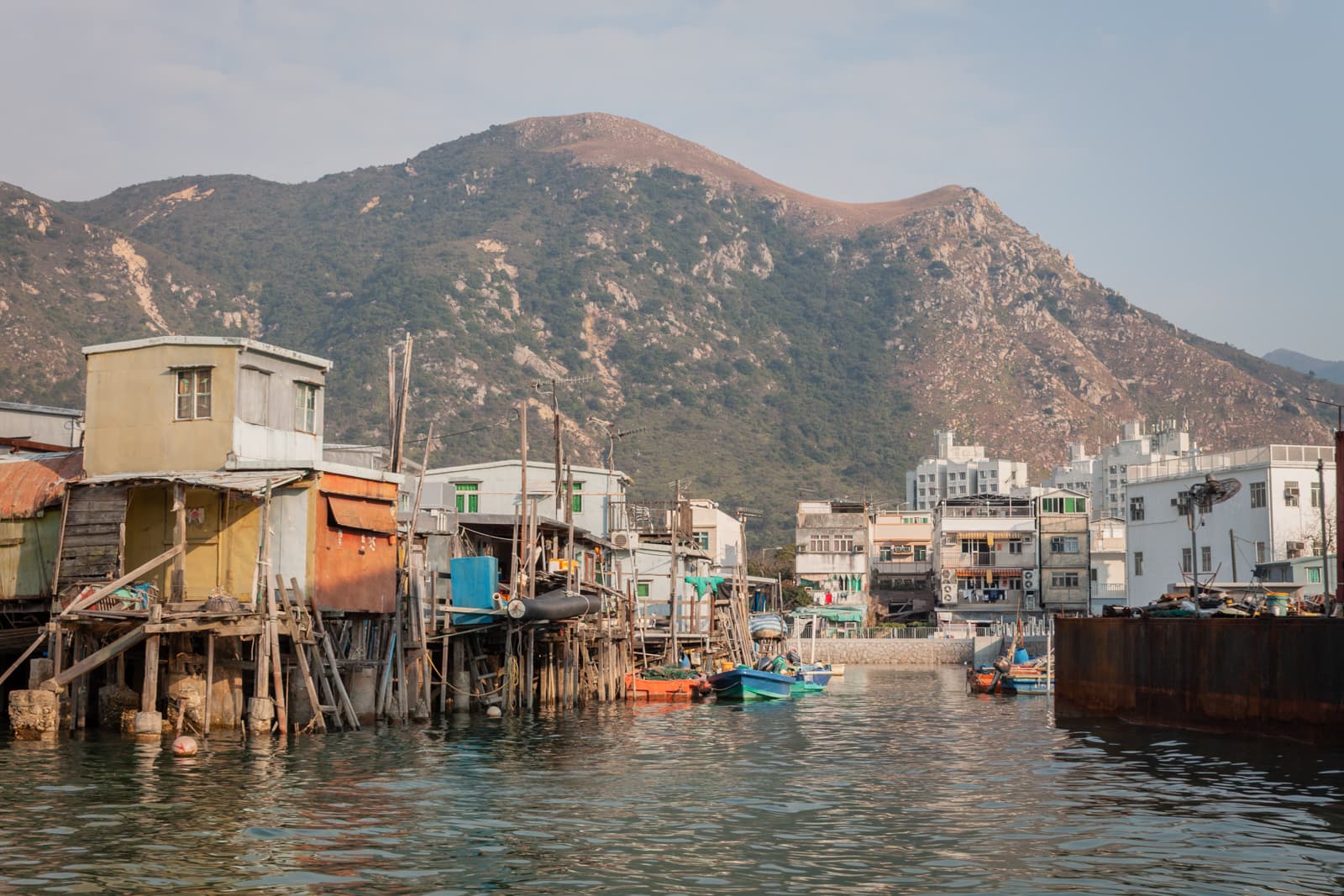 A rustic waterfront village with stilt houses rising above calm waters. Colorful fishing boats are docked along the edges, with a mountainous landscape providing a backdrop.