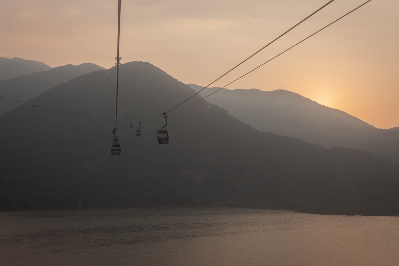 Cable cars gliding over a large body of water at sunset, with silhouetted mountains in the background. The fading sunlight creates a soft, golden hue across the sky and water.