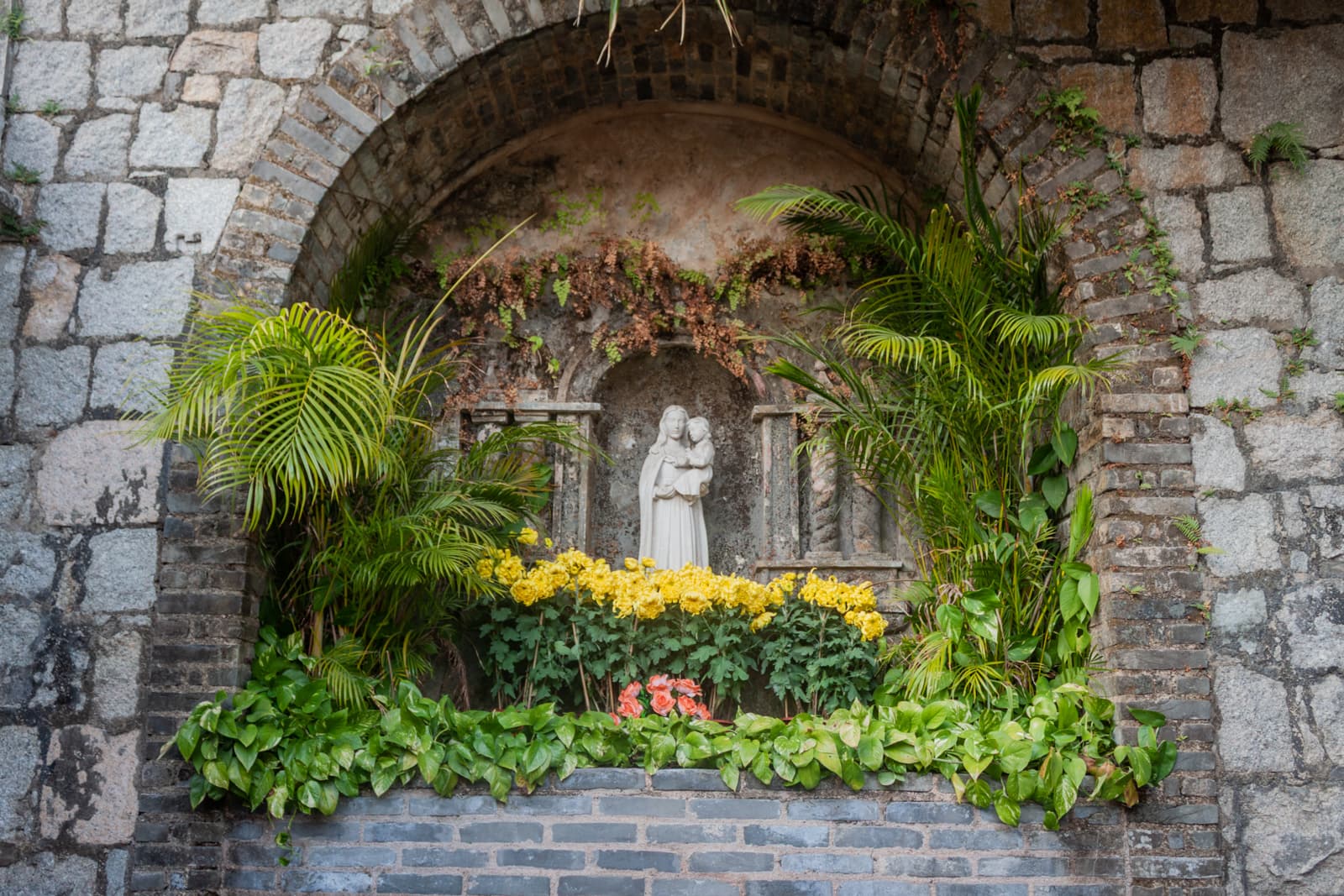 A serene alcove built into a stone wall, housing a statue of the Virgin Mary holding baby Jesus. The alcove is adorned with lush greenery, including palm fronds and vibrant yellow flowers.