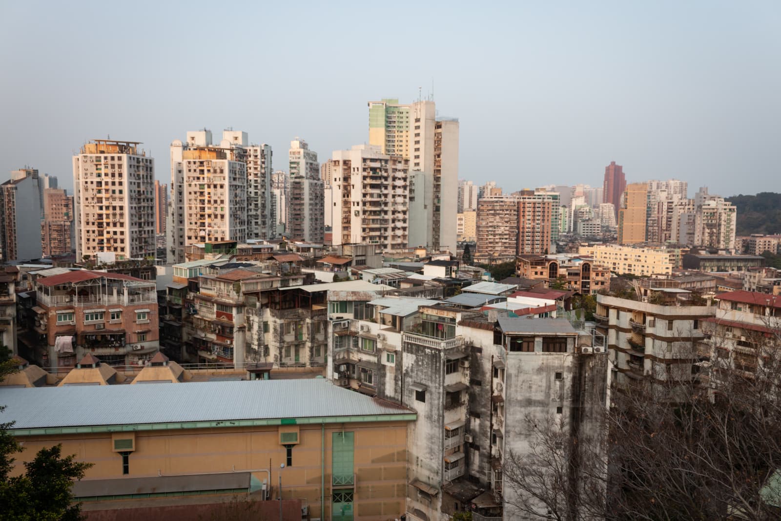 A sprawling cityscape featuring a mix of old, weathered buildings and modern high-rise apartment towers. The buildings are packed closely together, and the sky is clear with soft afternoon light.