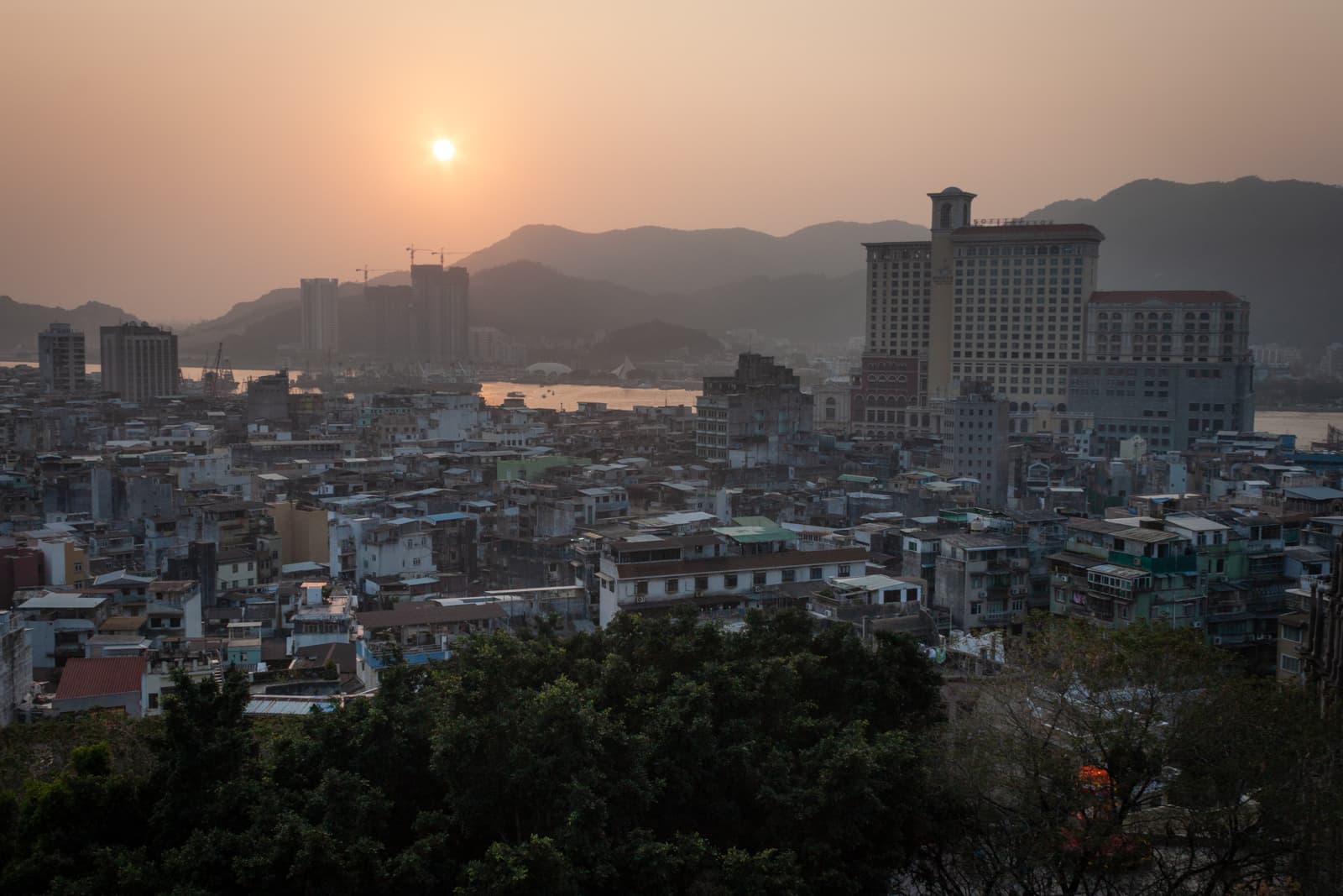 A hazy sunset over a densely packed urban landscape, with low-rise buildings in the foreground and taller structures in the distance. Mountains and a river can be seen beyond the city, softly lit by the setting sun.