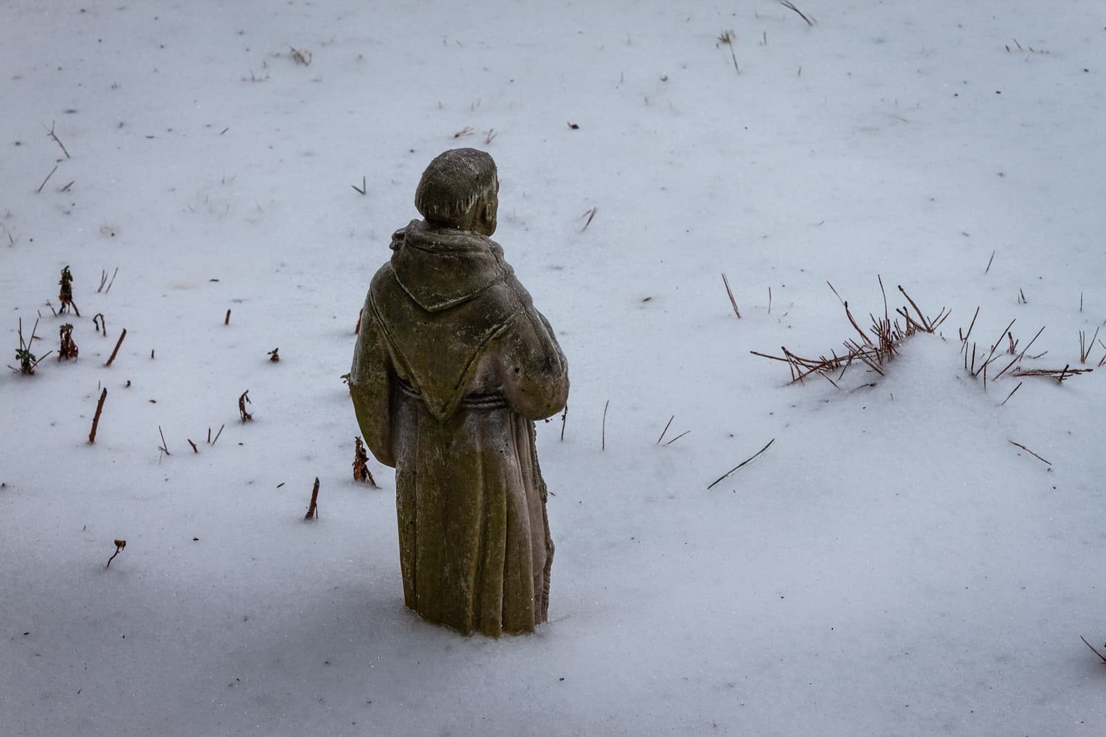 A small statue of a monk, half-buried in snow, standing in a serene, snow-covered field. The surrounding landscape is sparse with just a few tufts of grass peeking through the blanket of white.