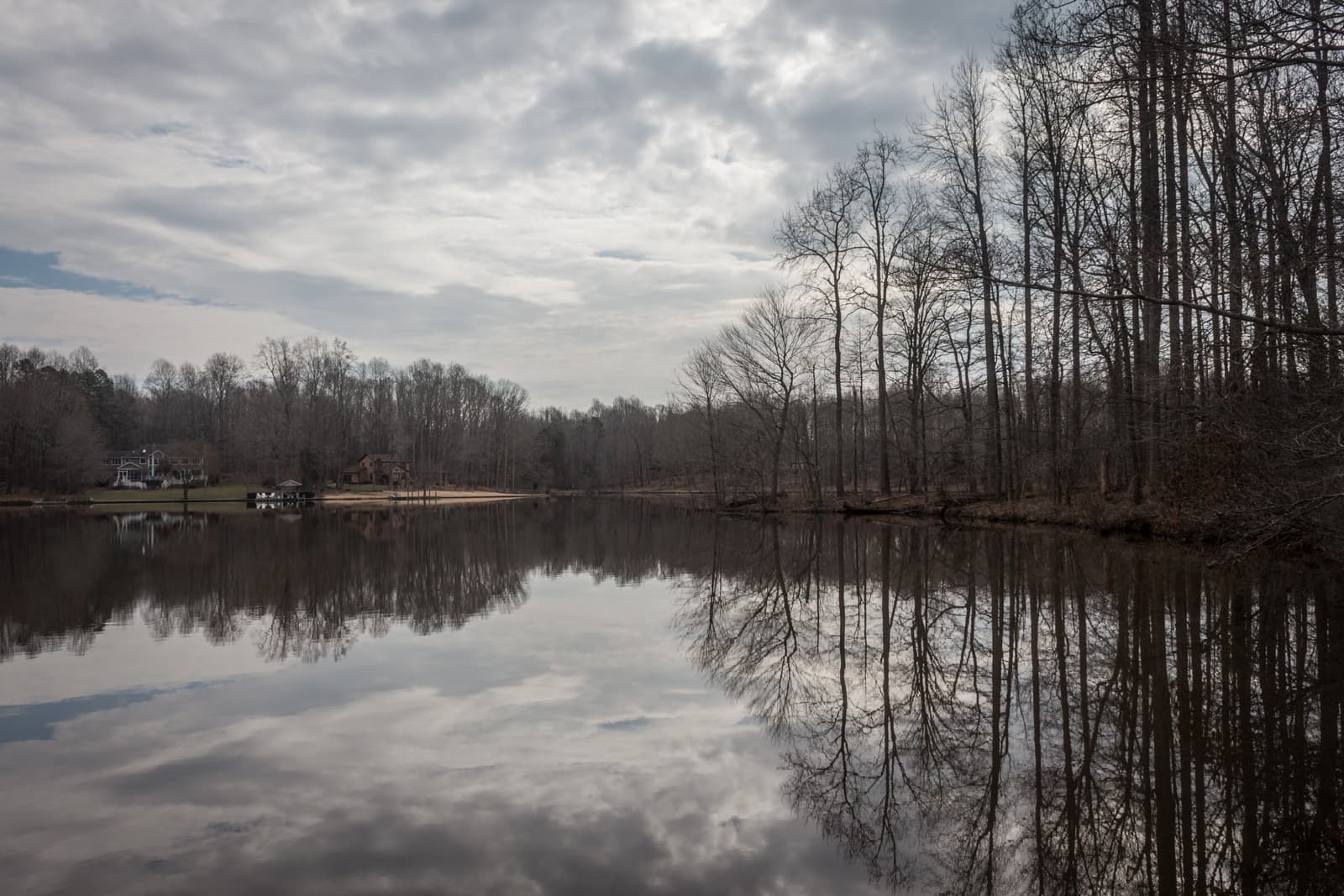 A calm lake reflects the overcast sky and the trees lining the shore. The still water mirrors the leafless trees, while houses and docks sit quietly along the far side of the lake, adding to the peaceful winter landscape.