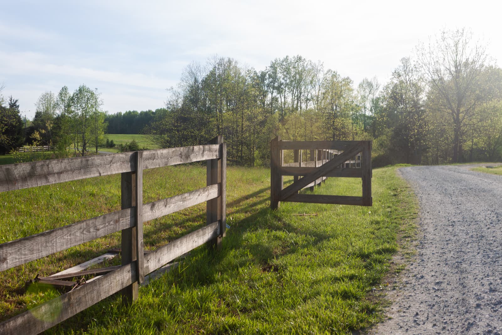A quiet country road with a wooden fence running alongside a grassy field. The road is gravel, and the scene is bathed in warm, late afternoon sunlight, creating a serene and inviting rural landscape.