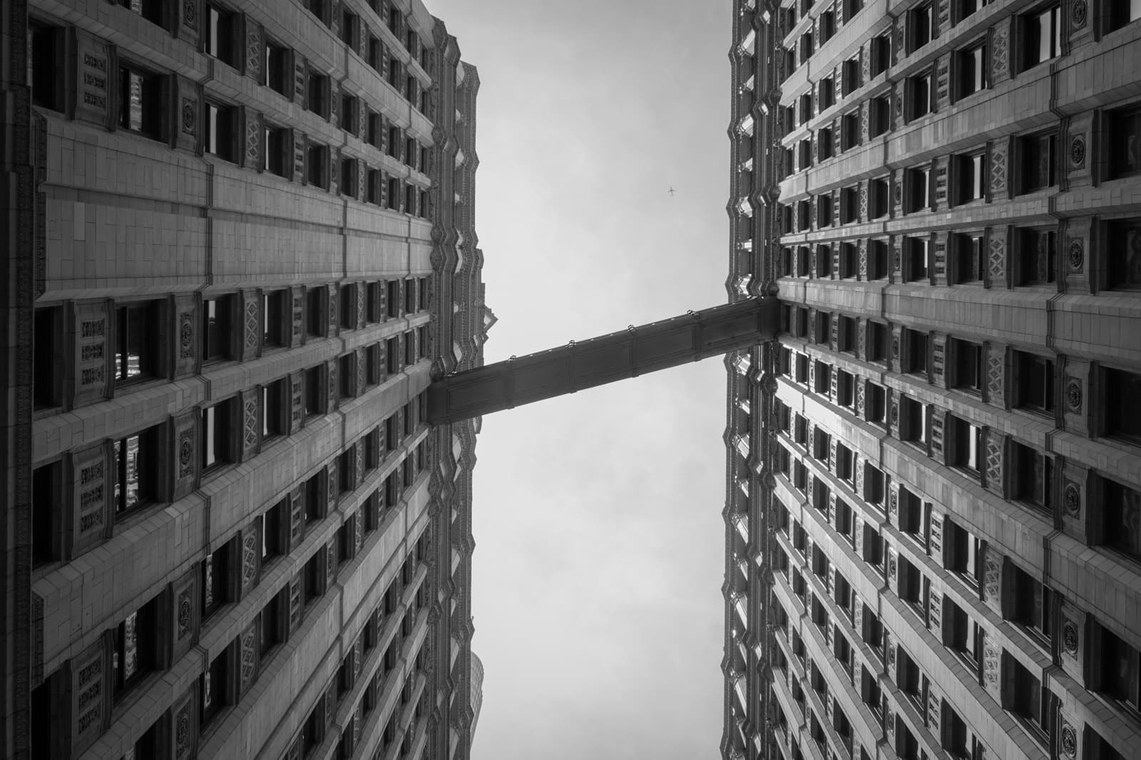 A black-and-white upward shot of two tall, symmetrical buildings with an enclosed bridge connecting them at a high level. The perspective creates a dramatic, towering effect as the buildings seem to converge toward the sky.