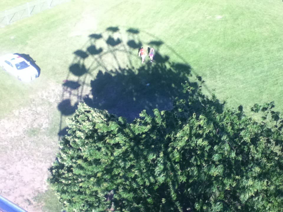 Aerial view of a grassy field with the shadow of a Ferris wheel cast on the ground. The shadow is partially obscured by a tree, with a few people standing in its shade. To the left, a parked car is visible. The bright sunlight and contrast create a playful scene, highlighting the Ferris wheel's circular form from an unseen vantage point.