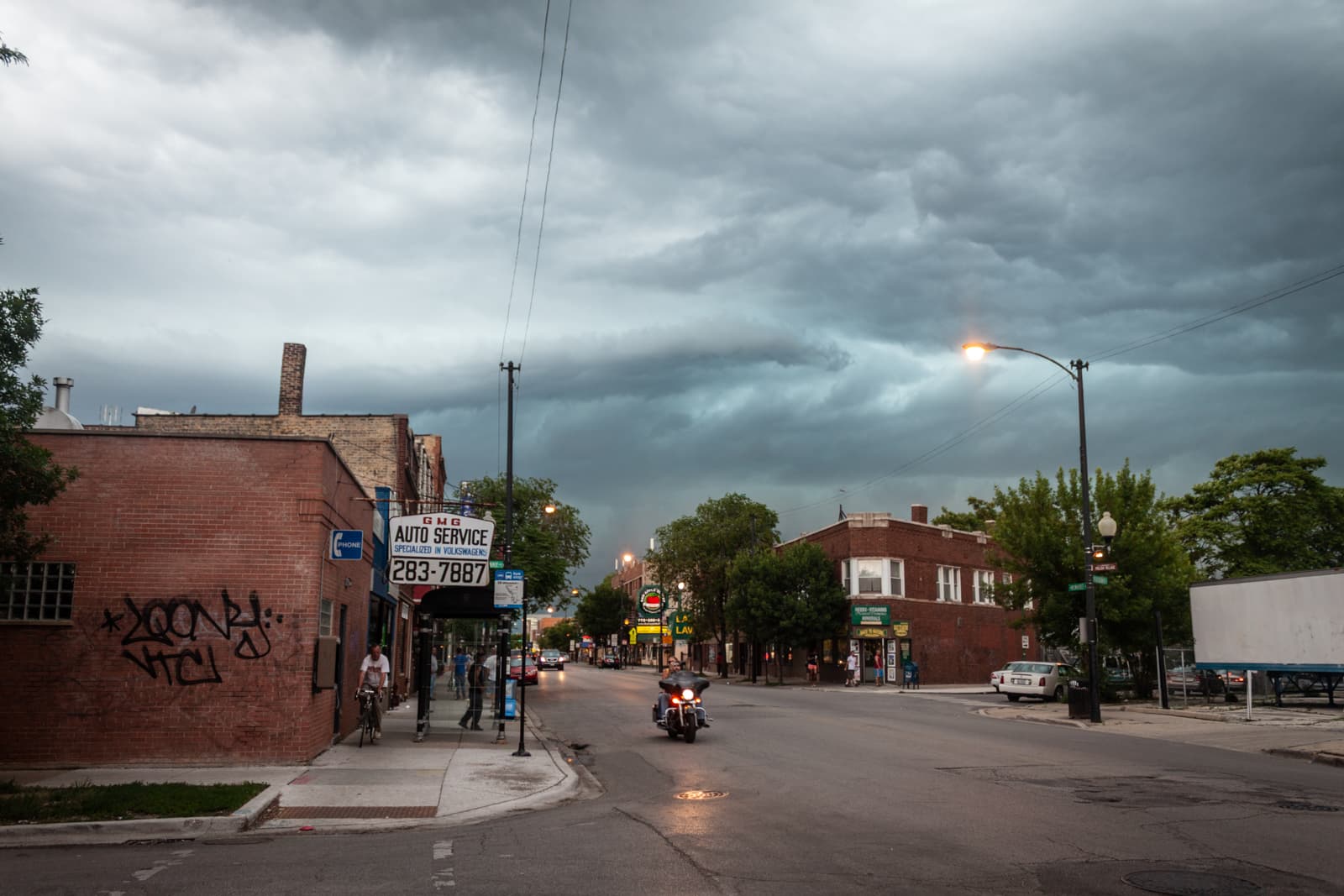 A wide-angle view of a quiet urban street under a dark, stormy sky. The street is lined with brick buildings, including an auto service shop with a prominent sign and graffiti on its side. A motorcyclist is seen riding down the road, while pedestrians walk on the sidewalk, giving a sense of calm before the impending storm. The ominous clouds contrast with the soft streetlights, creating a dramatic atmosphere.