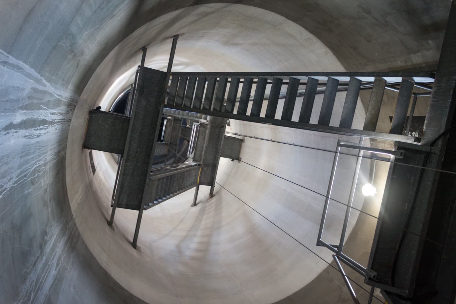 A steep, industrial staircase spirals upwards inside a large, cylindrical concrete shaft. The metal stairway is sharply angled, creating a dramatic perspective that draws the eye toward the distant opening at the top. The walls of the shaft are smooth, and faint streaks of water stains run along the surface, adding texture to the otherwise stark surroundings. The lighting is minimal, with a single bare bulb casting shadows and emphasizing the utilitarian design of the structure.