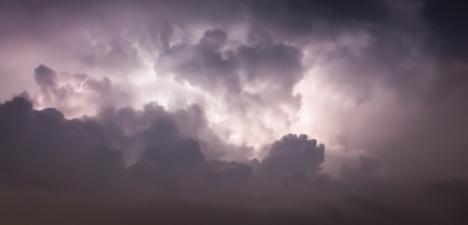 A dramatic scene of storm clouds illuminated by flashes of lightning. The thick, dark clouds are backlit, creating a glowing, ethereal effect with soft gradients of light and shadow. The faint outline of a lightning bolt can be seen streaking through the clouds, adding intensity to the otherwise calm, moody atmosphere. The sky is a blend of dark purples and grays, giving a sense of the power and beauty of a storm in motion.
