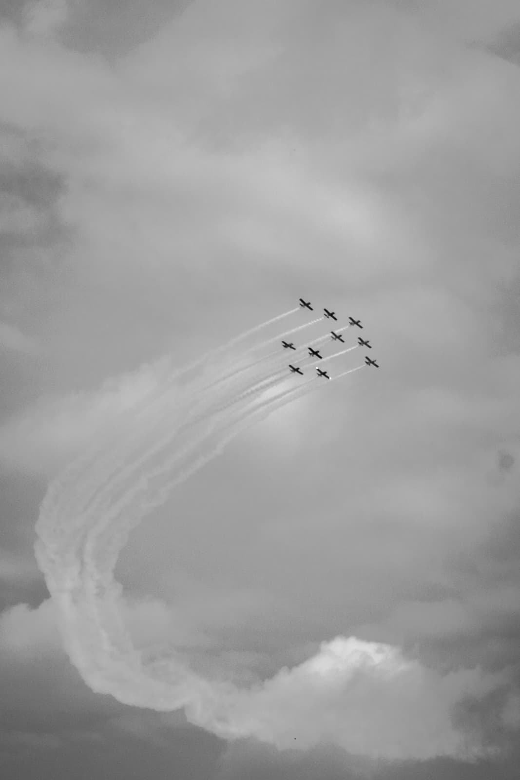 Seven airplanes flying in an arc-shaped formation, leaving smoke trails that curve behind them, depicted in black and white.