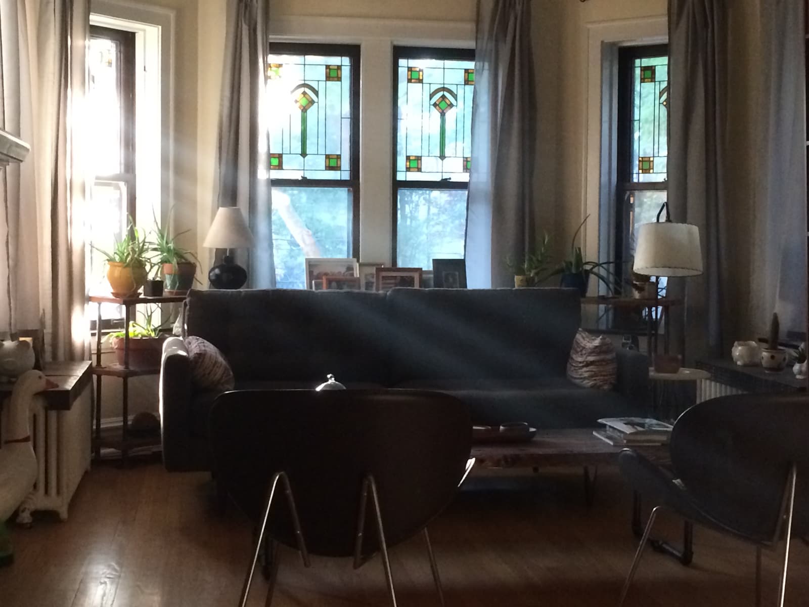 Cozy living room with sunlight streaming through stained glass windows. A dark couch is positioned against the windows, with a few plants on a side table and two lamps on either side. Wooden furniture and chairs complete the inviting atmosphere, with light filtering softly into the room.