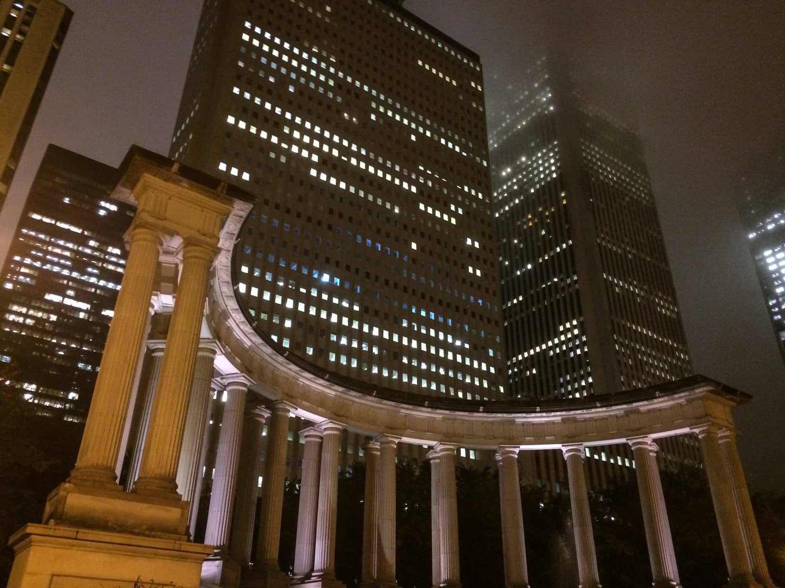 Nighttime view of a neoclassical colonnade lit from below, with tall, illuminated office buildings towering in the background. The tops of the skyscrapers fade into the foggy night sky, creating a contrast between the historic columns and modern architecture.