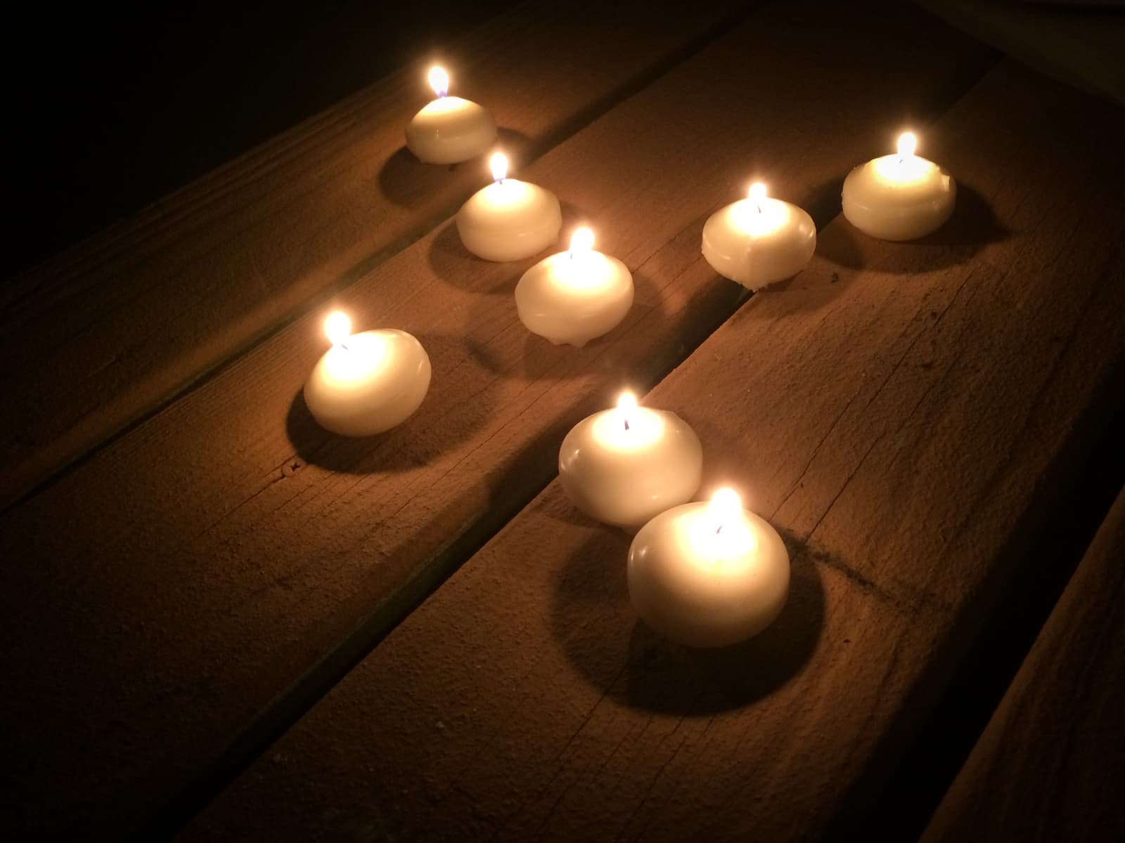 Several small tealight candles placed on a wooden surface, each softly glowing and casting gentle light and shadows in an otherwise dark environment.