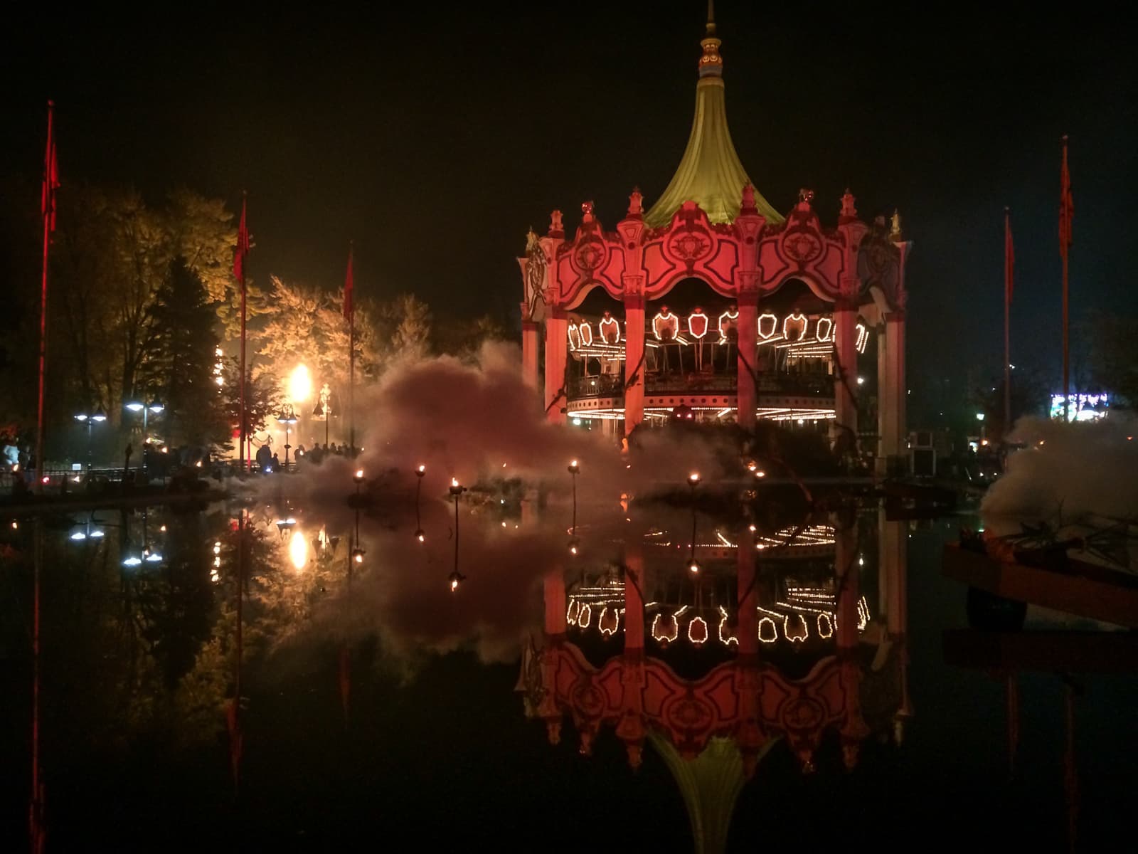 A grand carousel illuminated at night, with its reflection visible on the water in front. Surrounding the scene are smoke and fire effects, creating a dramatic atmosphere with trees and lights in the background.