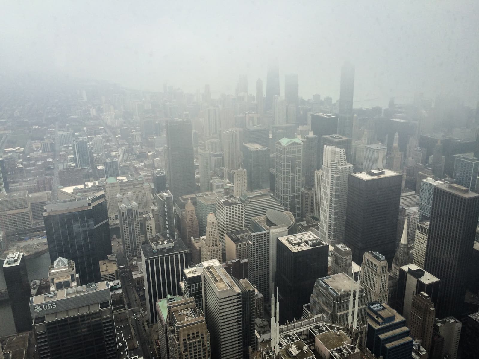 Aerial view of a fog-covered city skyline, with tall skyscrapers stretching out into the distance. The tops of the buildings and parts of the landscape are obscured by thick fog, creating a hazy, atmospheric scene.