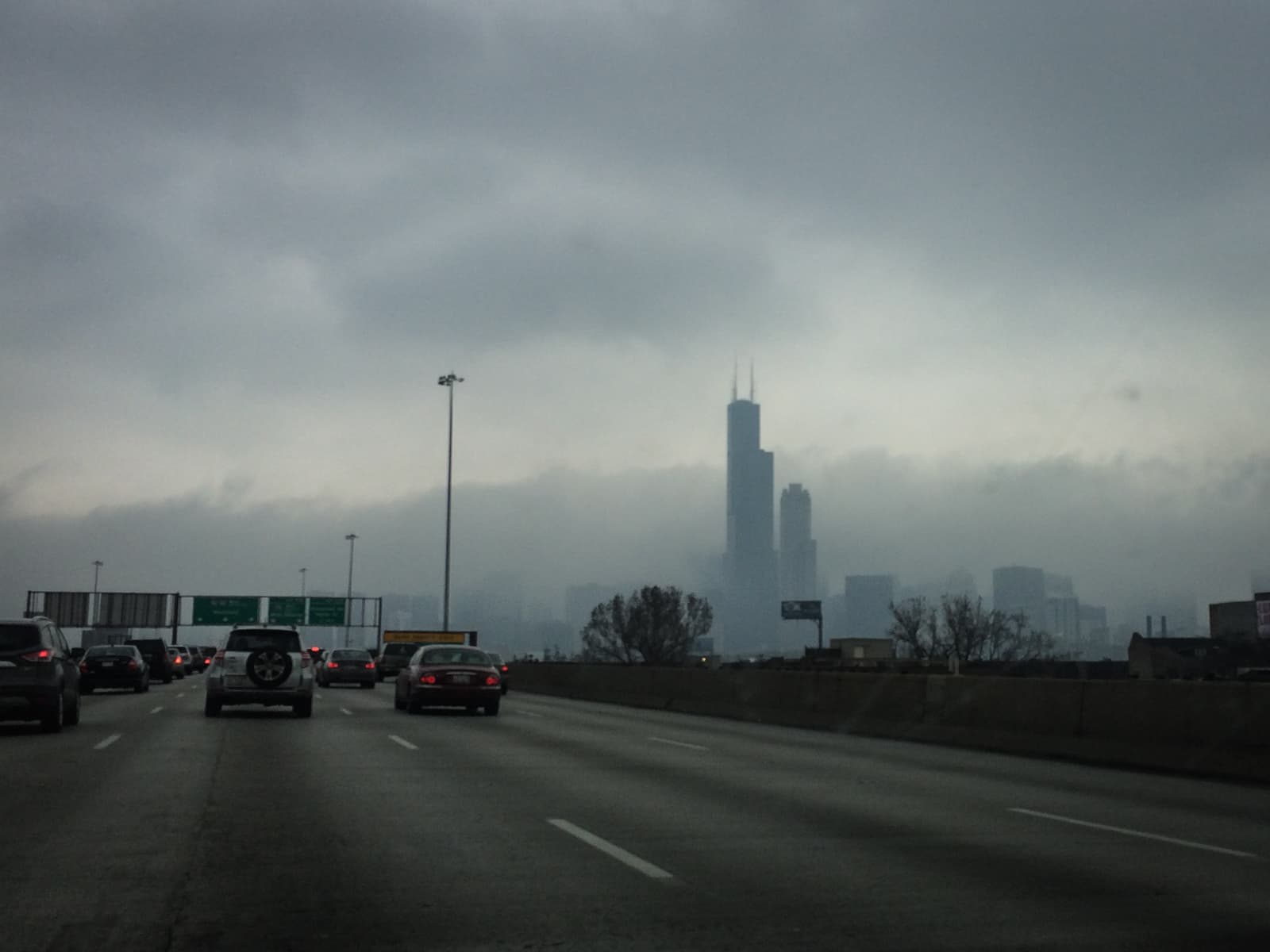 View from a car on a highway heading toward a fog-covered city skyline, with the silhouette of a tall skyscraper, the Willis Tower, faintly visible through the mist. The sky is overcast, and the road is lined with other vehicles.