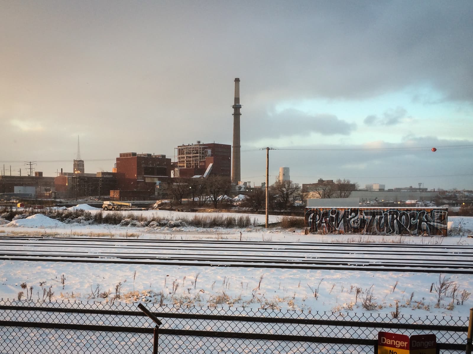 Industrial area in winter, with snow-covered ground and train tracks in the foreground. In the background are red brick buildings and a tall smokestack, with a large graffiti-covered wall near the tracks. The sky is partly cloudy, with a mix of warm and cool tones from the setting sun.
