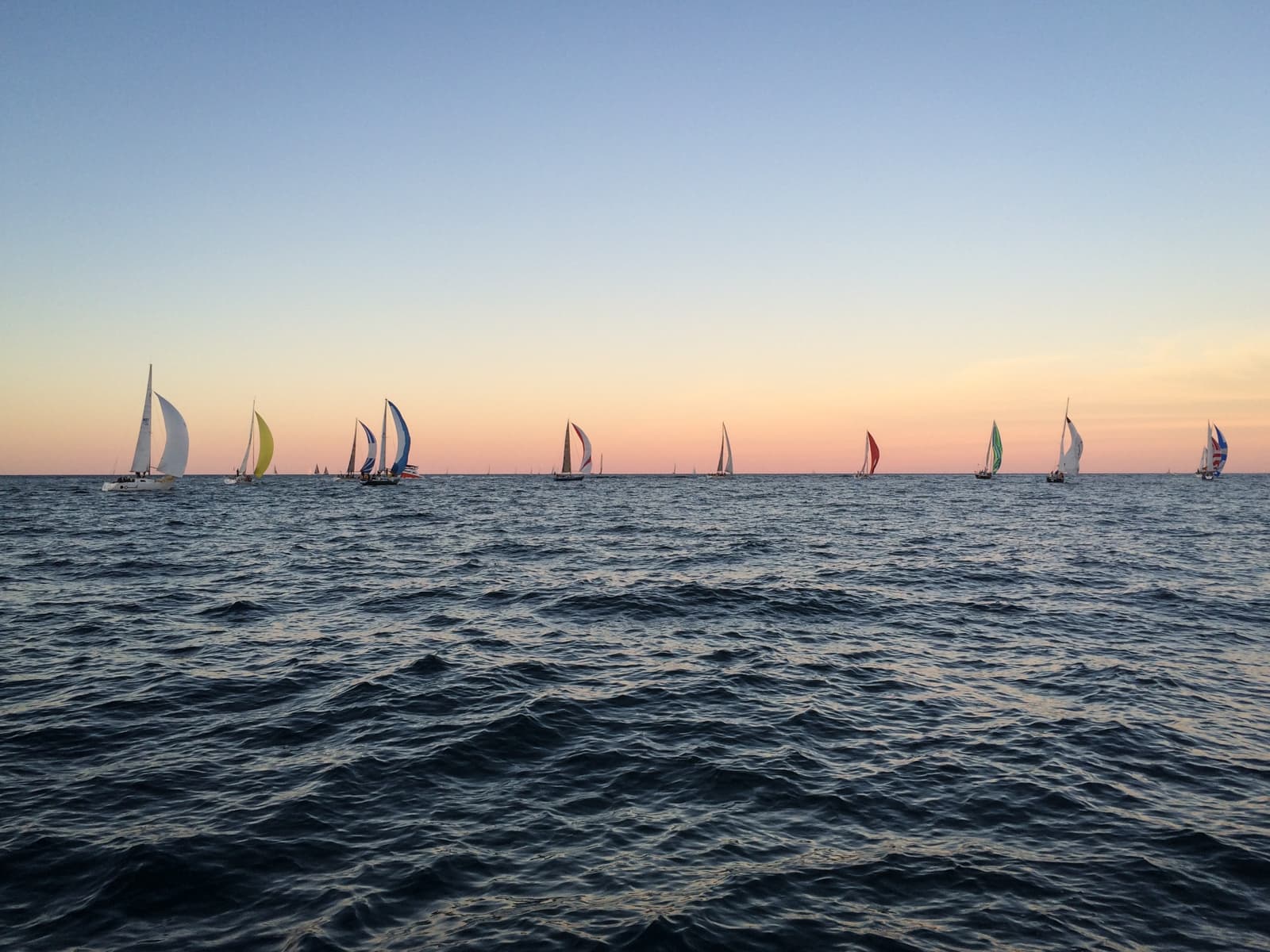 Several sailboats with colorful sails scattered across the water at sunset. The sky is a gradient of soft pinks, oranges, and blues, while gentle waves ripple across the surface of the sea.