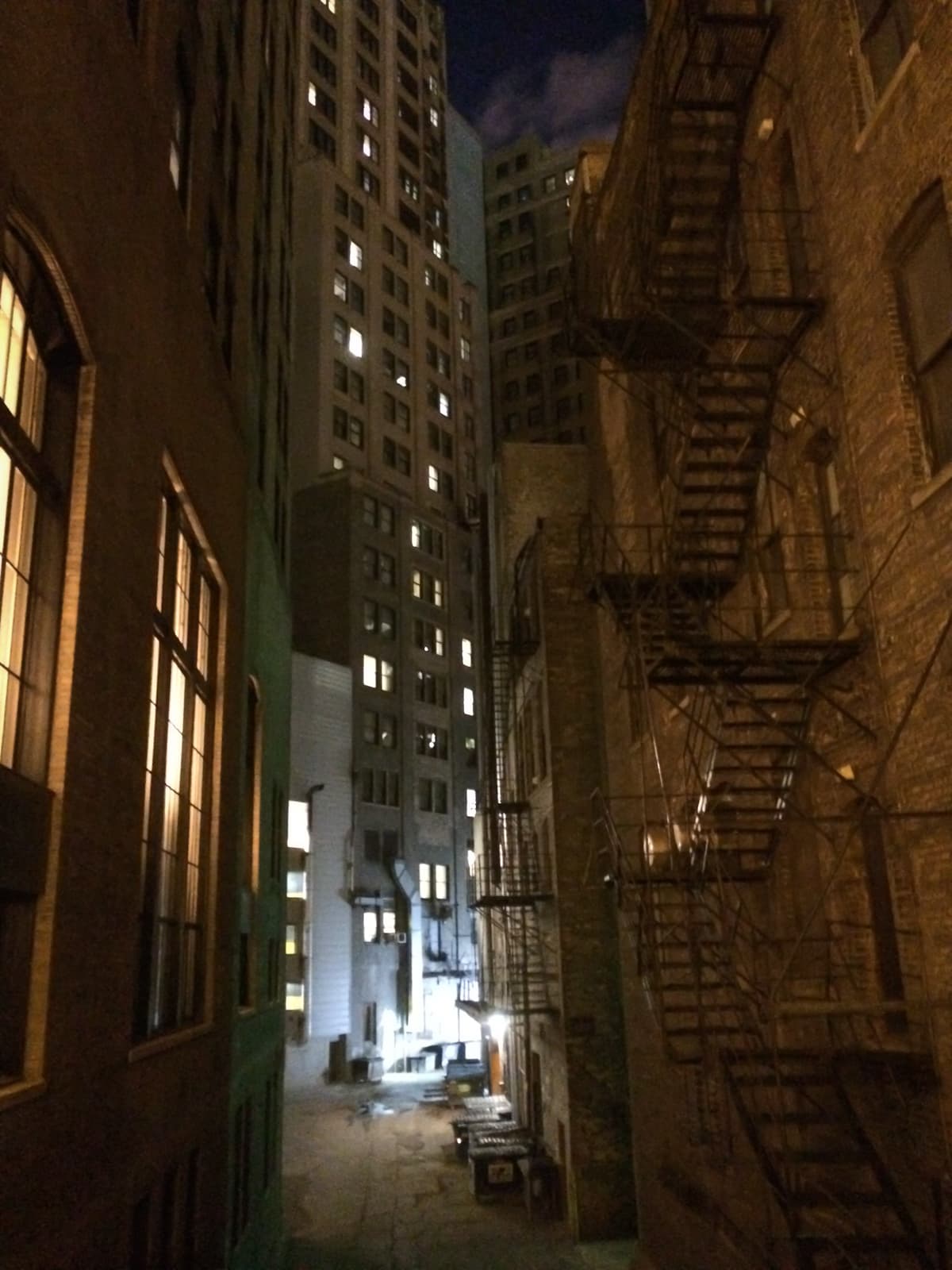 Dimly lit urban alleyway at night, surrounded by tall buildings with fire escapes on one side and windows glowing with light. The alley is empty, with trash bins near the far end, and the tops of skyscrapers rise into the night sky.