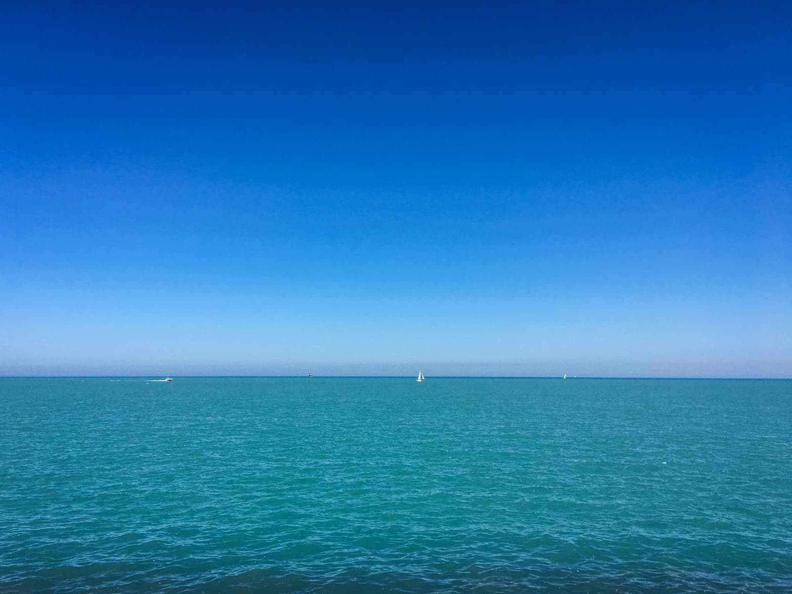 Clear, bright day over a calm turquoise sea, with a deep blue sky stretching above. A few small sailboats and distant vessels can be seen on the horizon, creating a peaceful and serene scene.