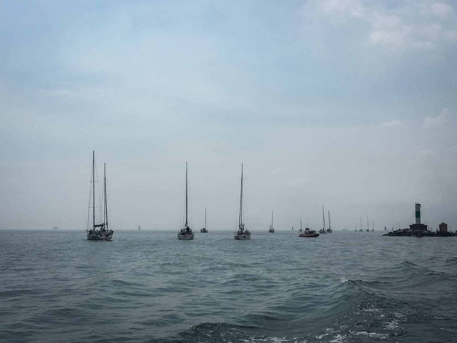 Several sailboats lined up in the distance on a calm sea, heading toward the horizon. A small lighthouse on the right marks the edge of a breakwater, while the sky remains overcast.