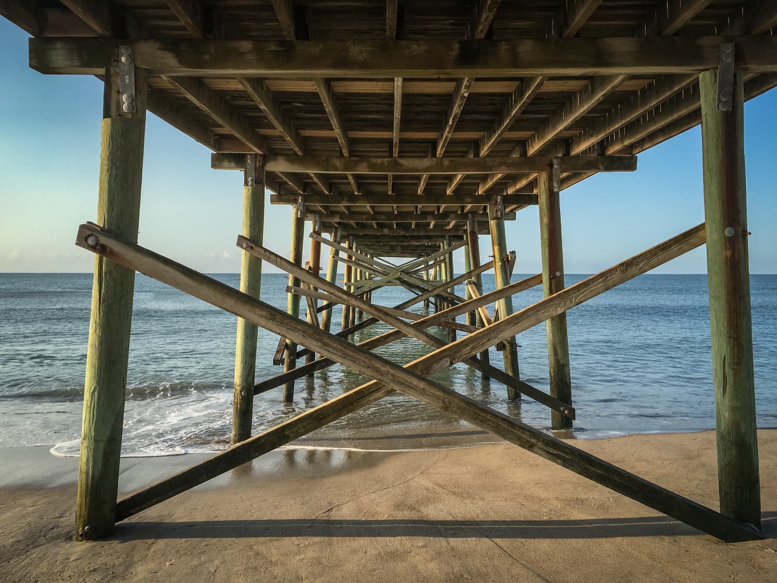 View from underneath a wooden pier stretching over the ocean, with diagonal support beams forming an ‘X’ pattern. The sandy beach and gentle waves are visible in the background, while the clear blue sky adds to the serene coastal scene.