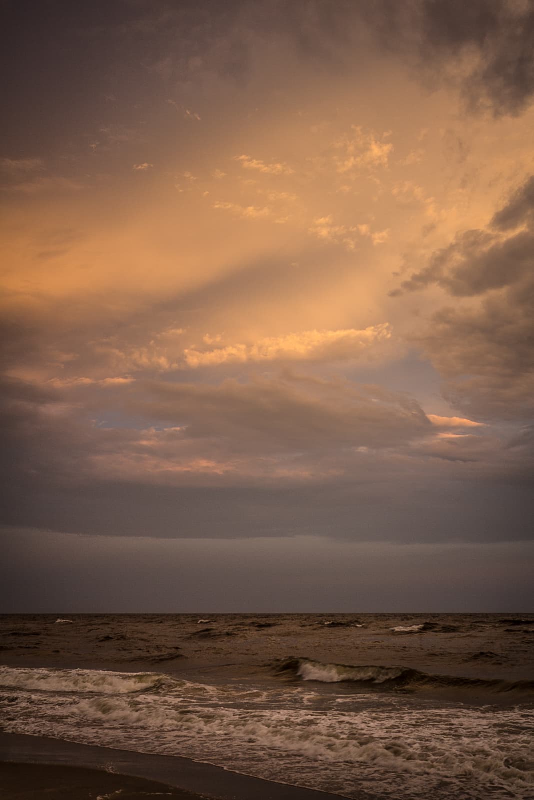 Dramatic seascape at dusk with waves crashing on the shore and a sky filled with layered, glowing clouds in shades of orange, gold, and purple.