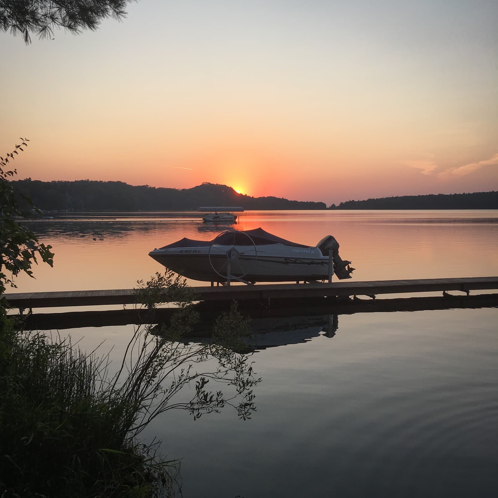 A peaceful lake at sunset, with the sun dipping behind distant hills and casting an orange glow over the water. A boat is docked at a wooden pier in the foreground, and the calm water reflects the colors of the sky.