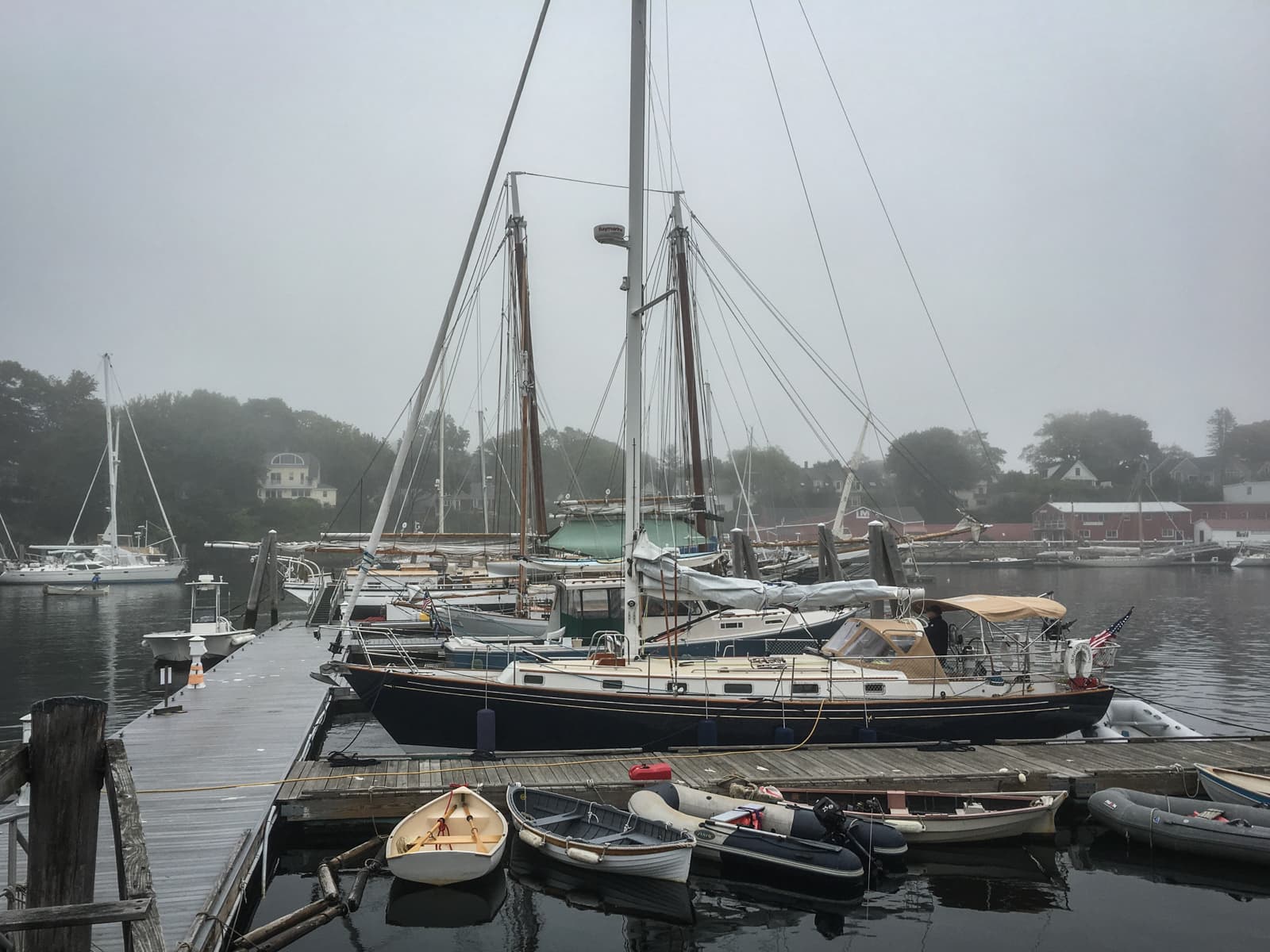 A foggy marina scene with several sailboats docked along a pier. Smaller boats are tied up in the foreground, while the distant shoreline and houses are barely visible through the mist. The atmosphere is quiet and calm, with muted tones.